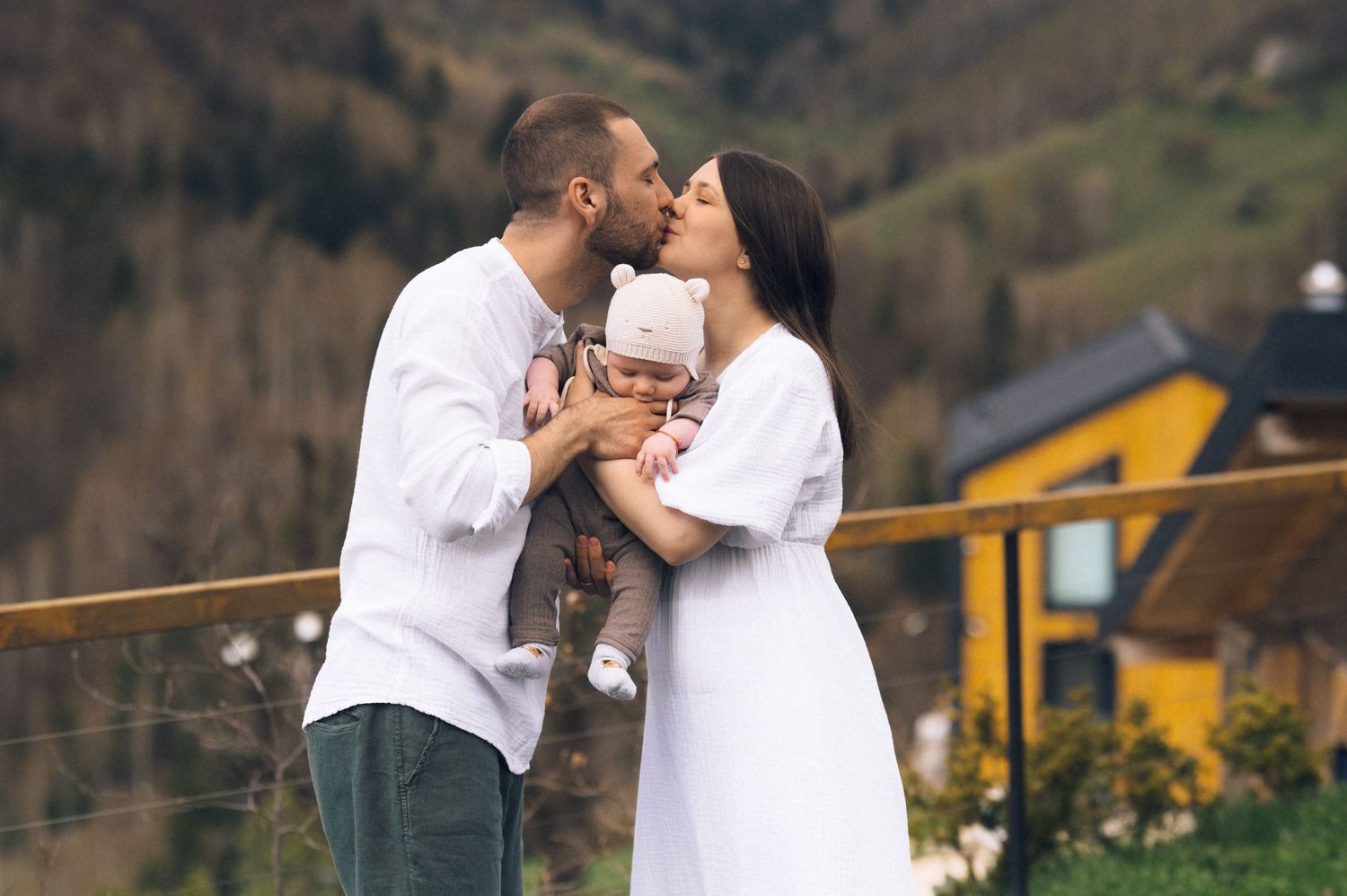 Young couple kissing while holding their newborn baby on a balcony with green mountain scenery behind.