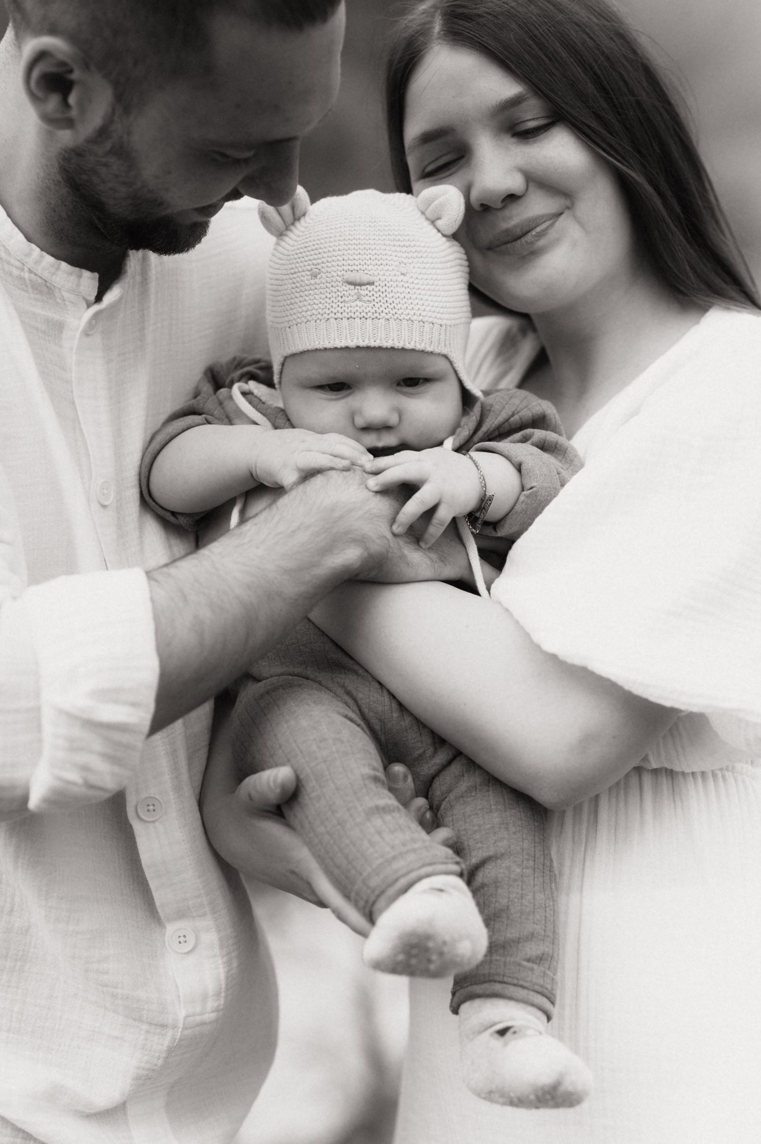 Black and white photo of parents lovingly holding their baby in a knit bear hat, capturing a tender family moment.