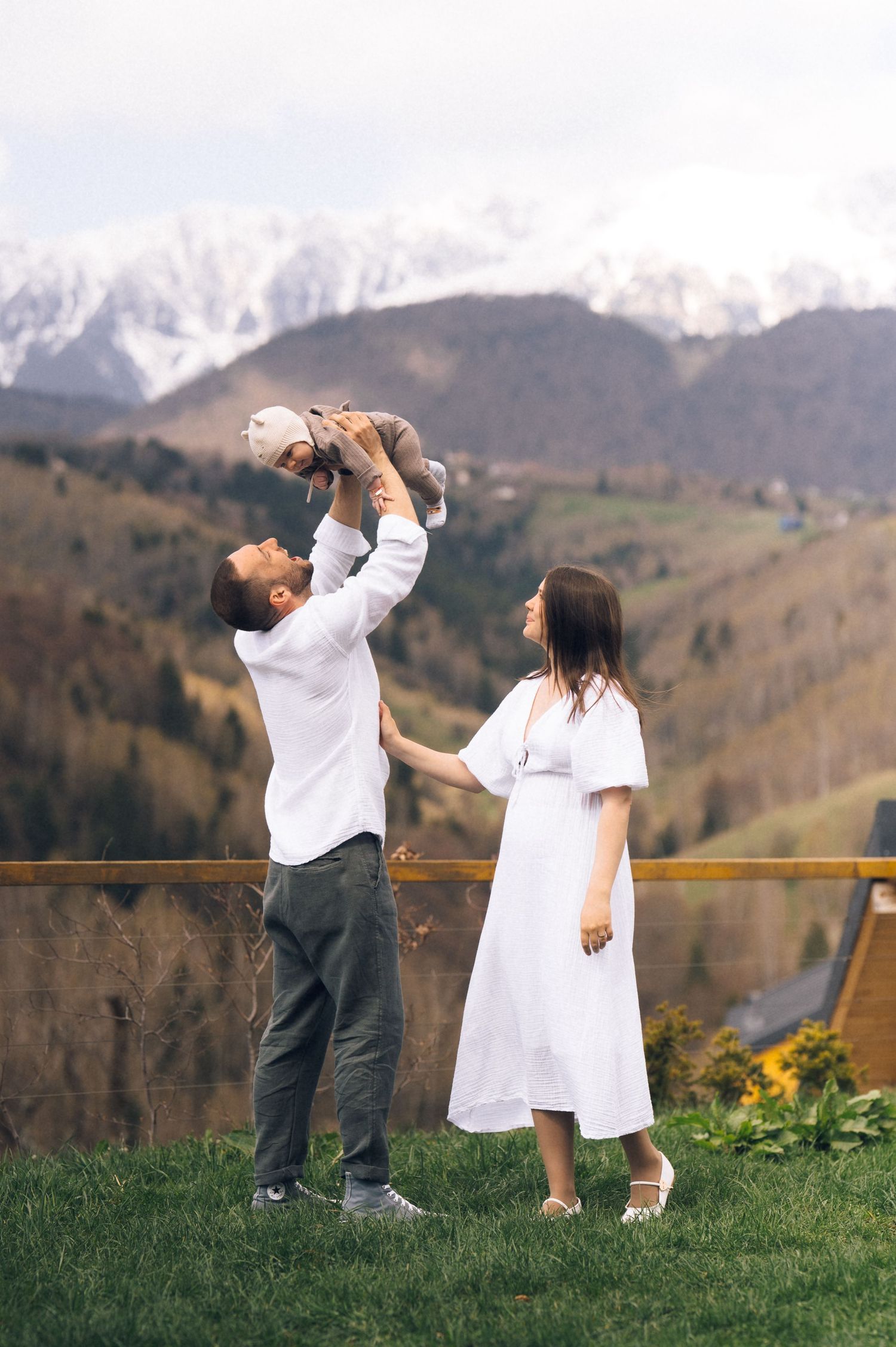 A joyful family with a man lifting a baby high while a woman in white dress stands beside, set against snowy mountains.
