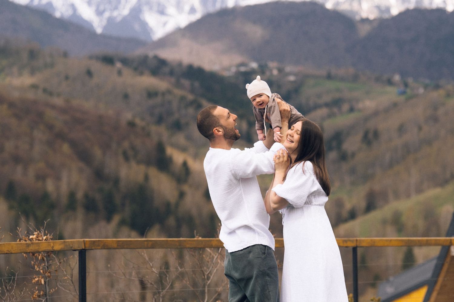 A couple holds their baby up against a scenic mountain backdrop with snow-capped peaks and autumn foliage.