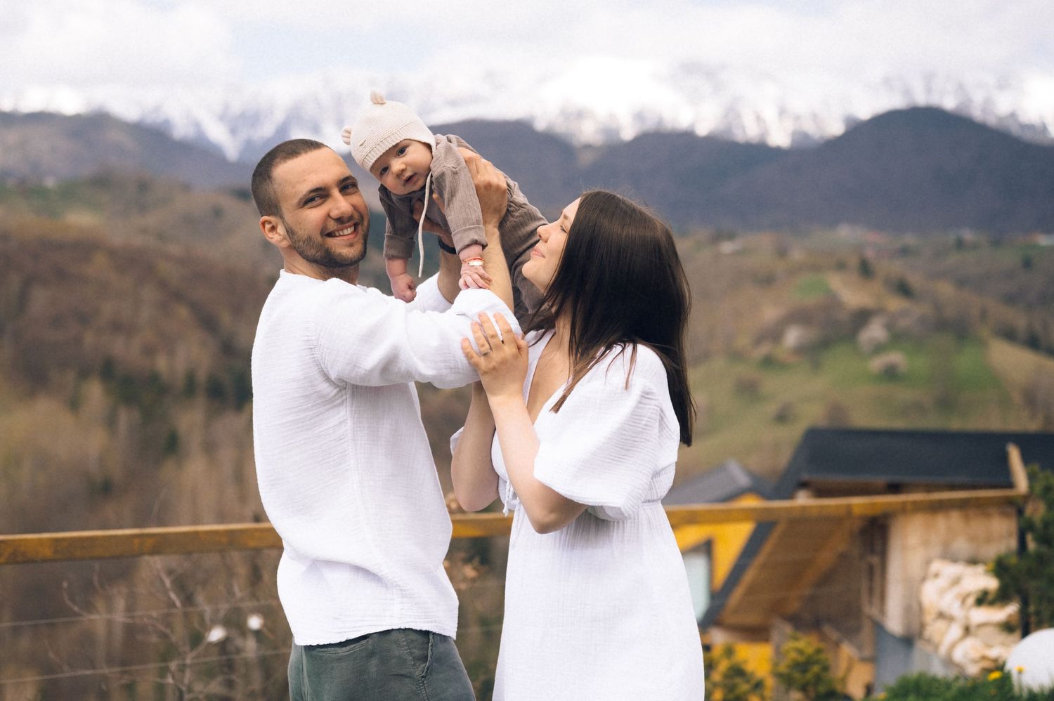 Happy family with baby enjoying mountain views, parents in white outfits lifting child outdoors.