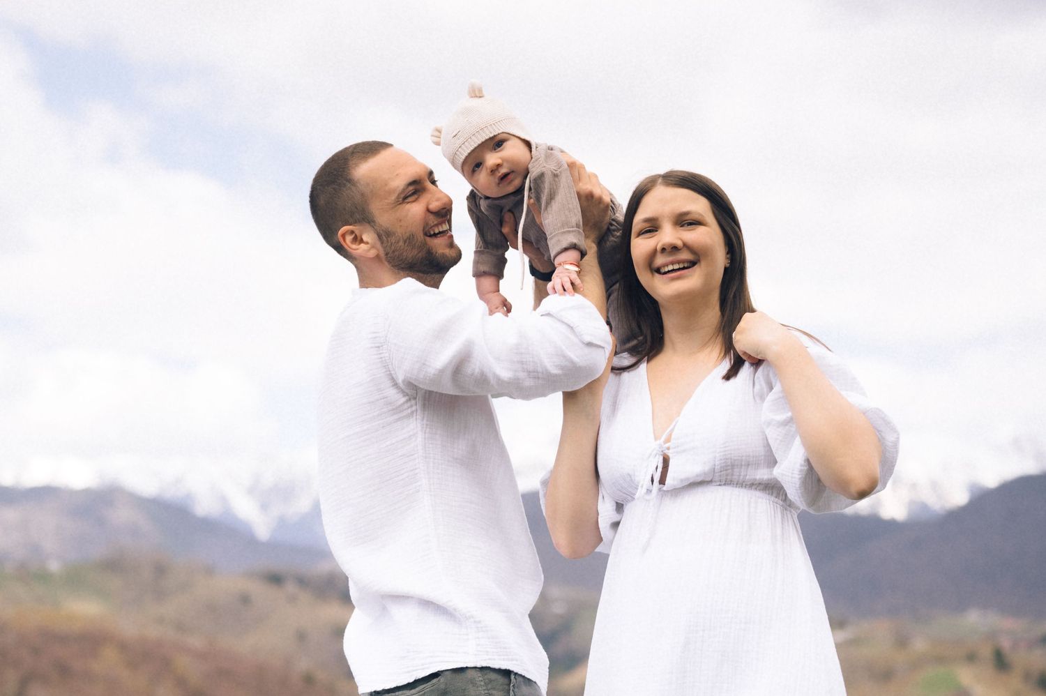 Happy family with father lifting baby girl, mother smiling, posing outdoors with mountain backdrop.