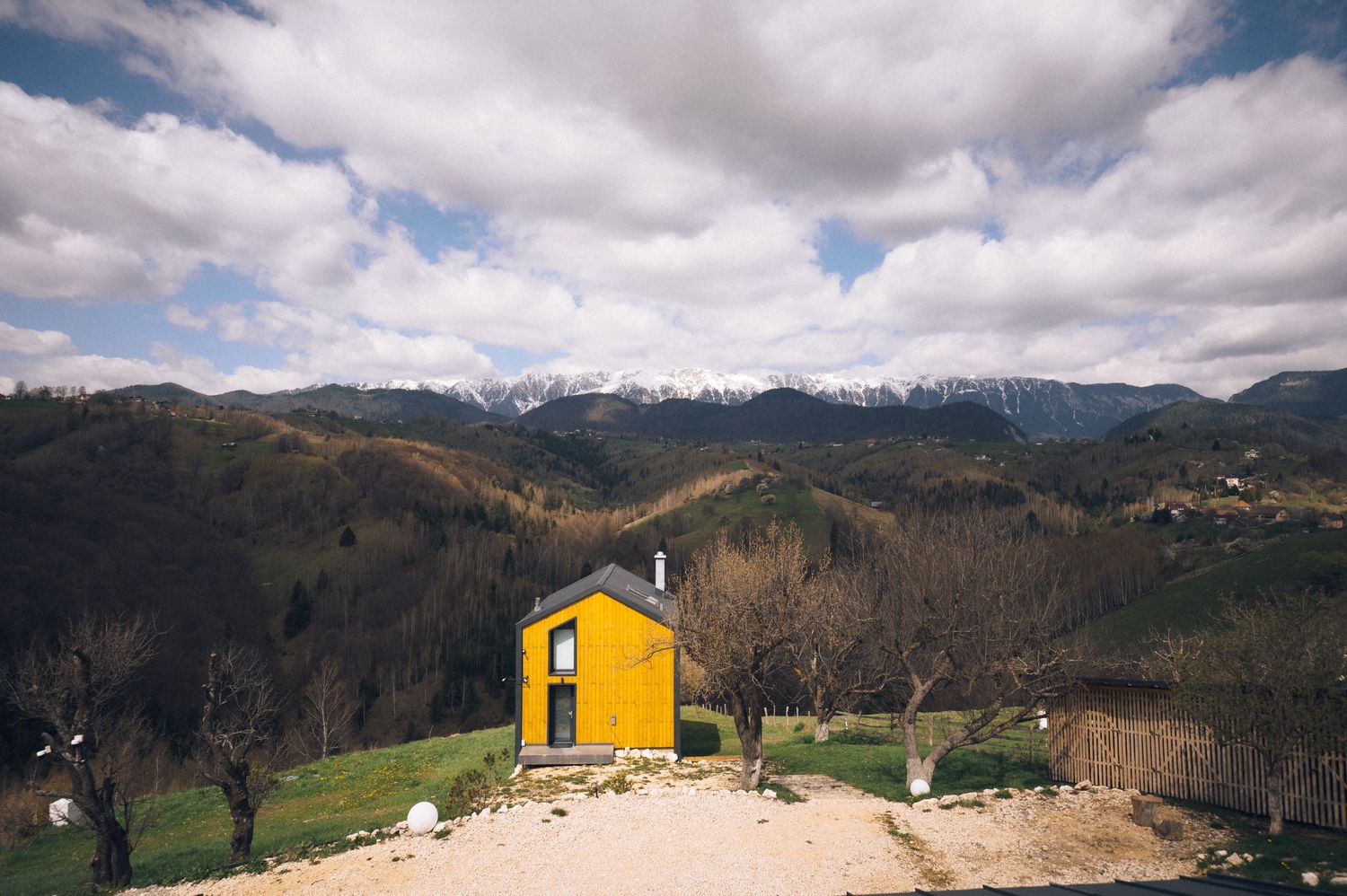 Bright yellow tiny house on a hillside with snow-capped mountains and cloudy sky in the background.