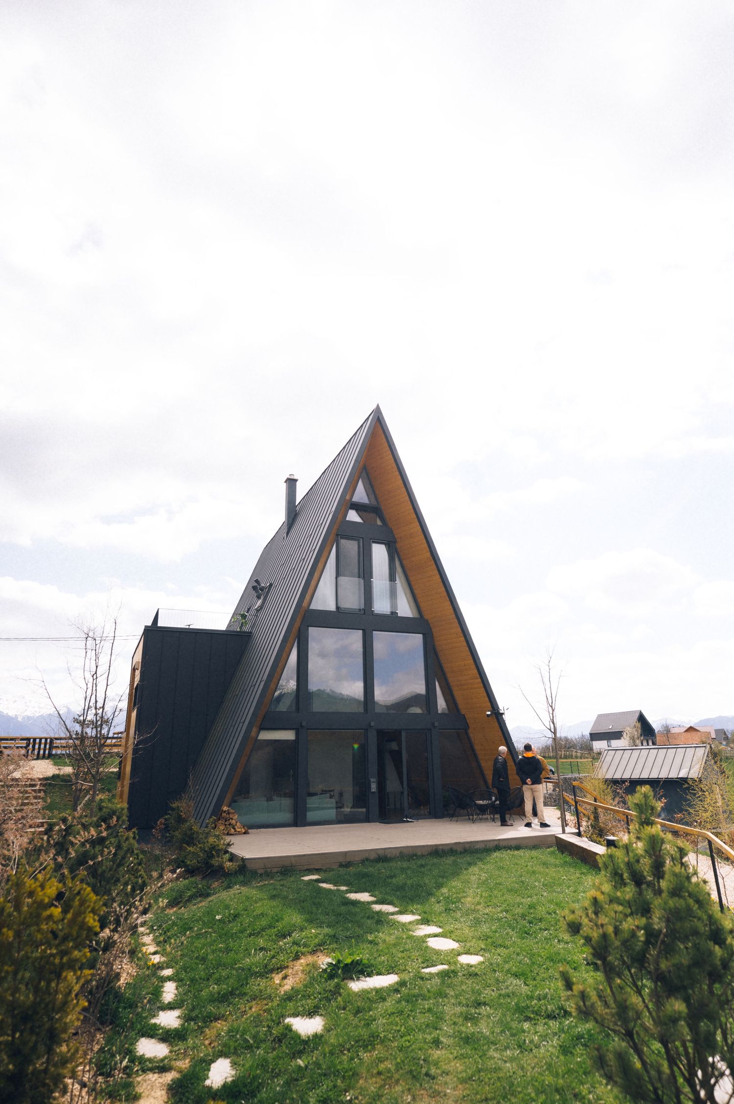 Modern A-frame cabin with black exterior, large glass windows, wooden deck, and lush green lawn under overcast skies.