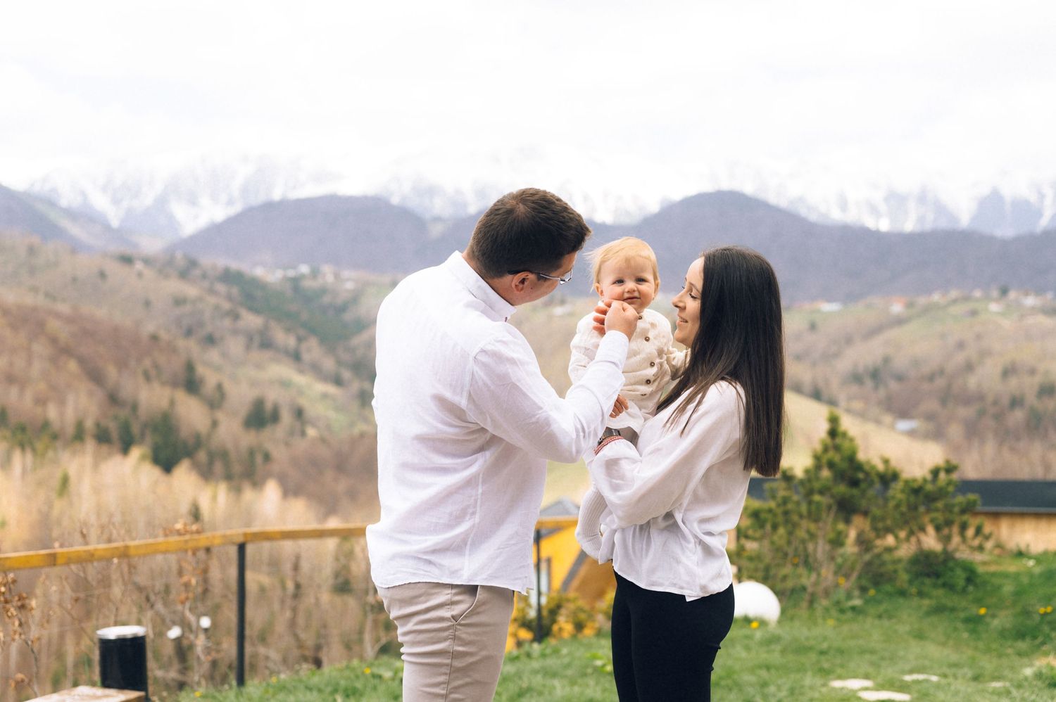 Young couple in white shirts holding their baby outdoors with scenic mountain landscape in background.