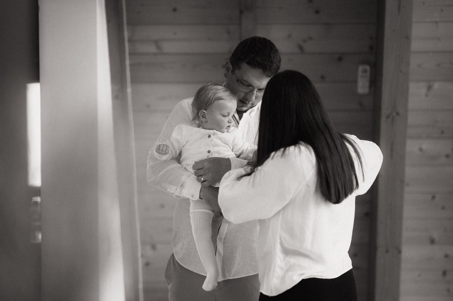 Black and white photo of a man holding a baby while a woman embraces them in a warm family moment.