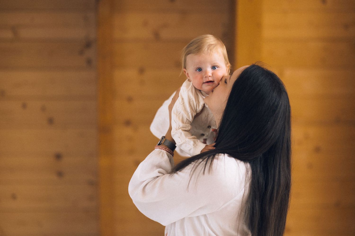 A mother in white holds her smiling baby up in the air inside a wooden room.