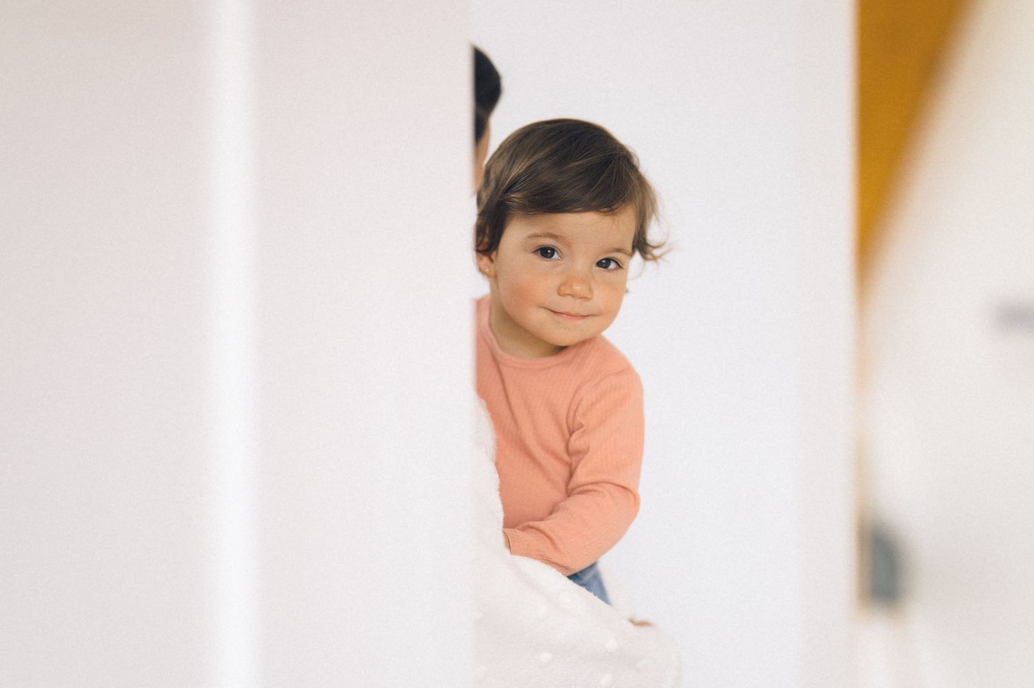 Cute toddler in a pink shirt peeking around a white wall with a curious expression.