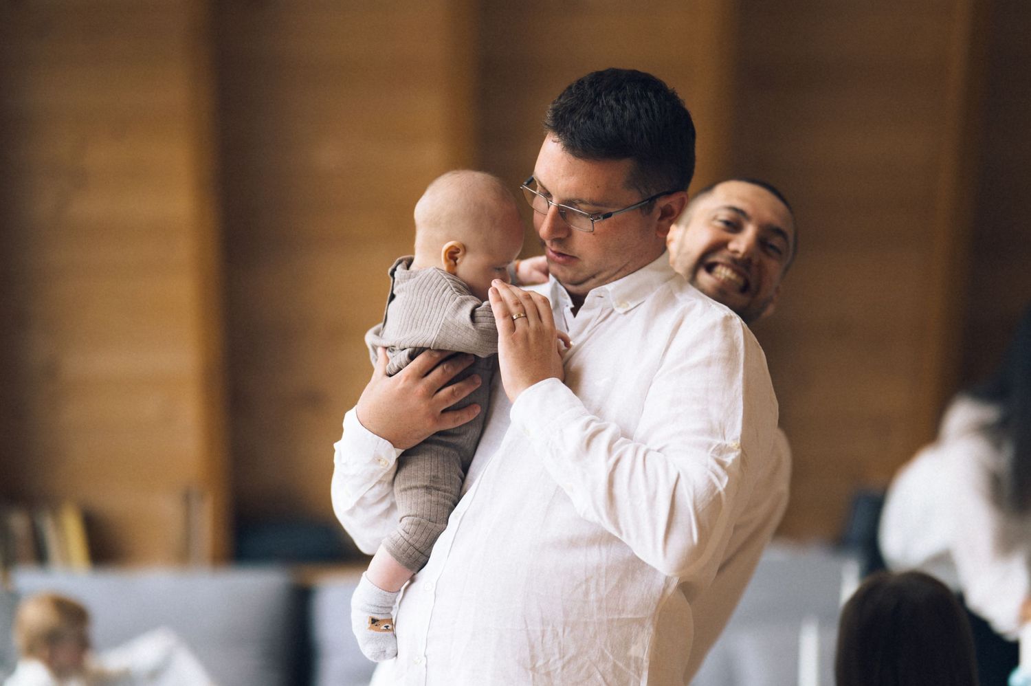 Man in white shirt tenderly holds a baby at an indoor event while another man smiles warmly in the background.
