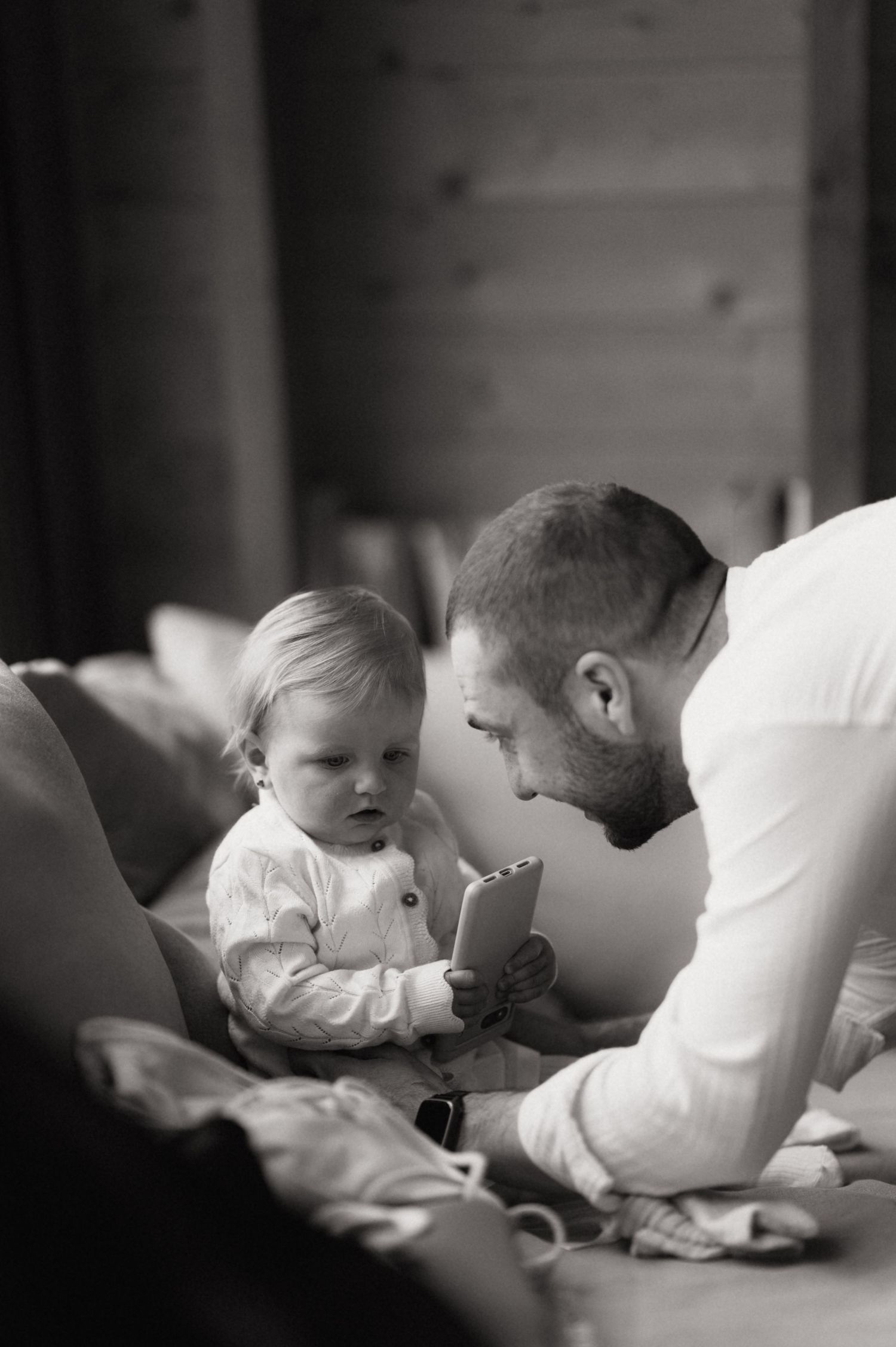 Black and white photo of a bearded father leaning in to interact with his toddler holding a small device on a bed.