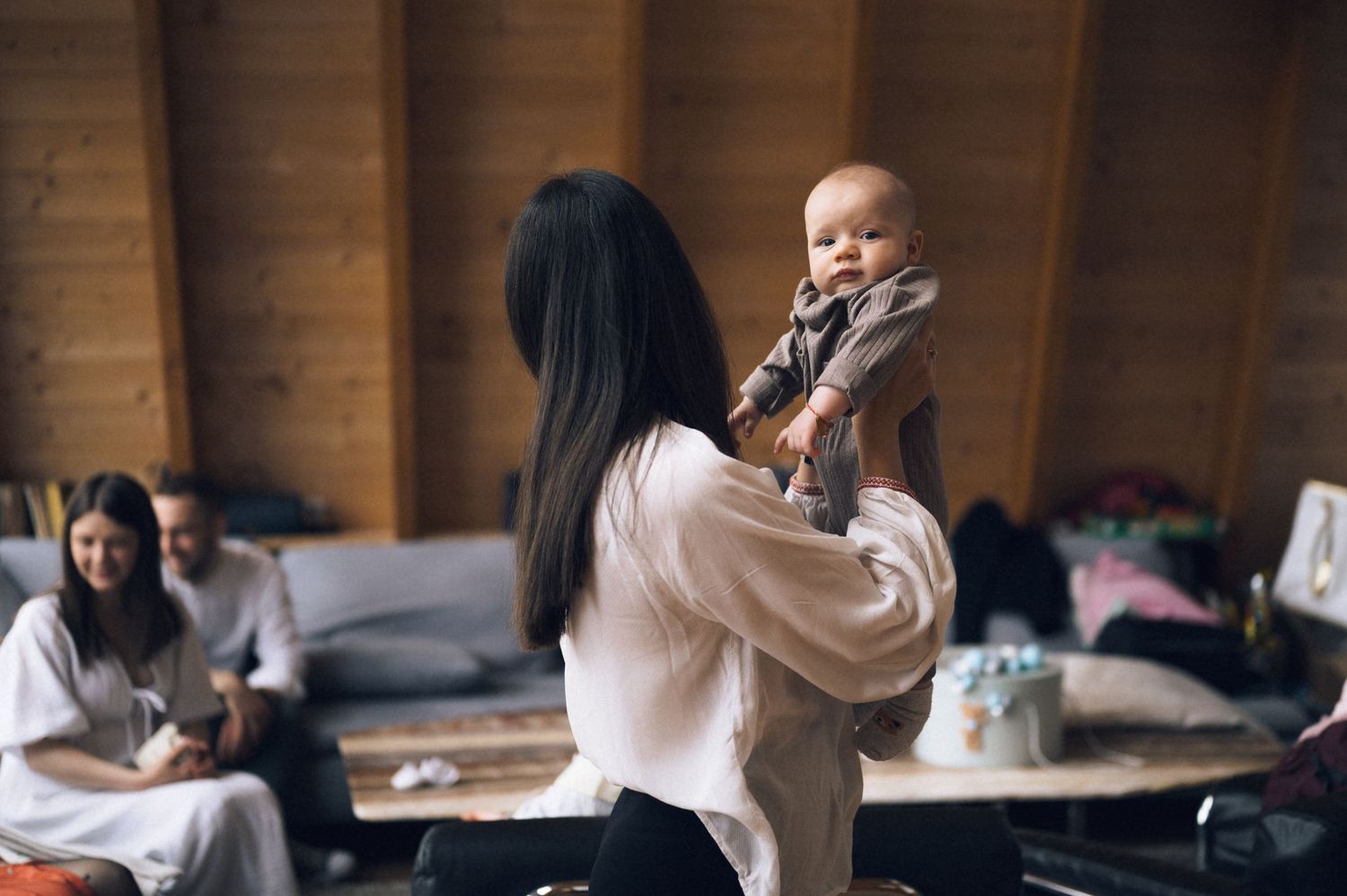 A woman holds a baby up in a warm wooden room while others sit in the background.