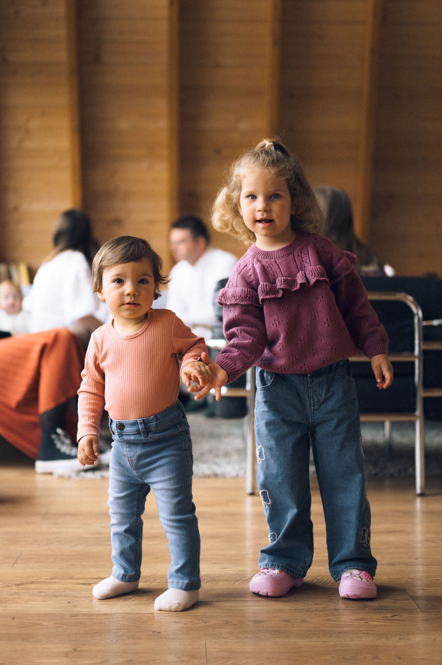 Two toddlers in matching jeans and colorful sweaters hold hands while standing on a wooden floor indoors.