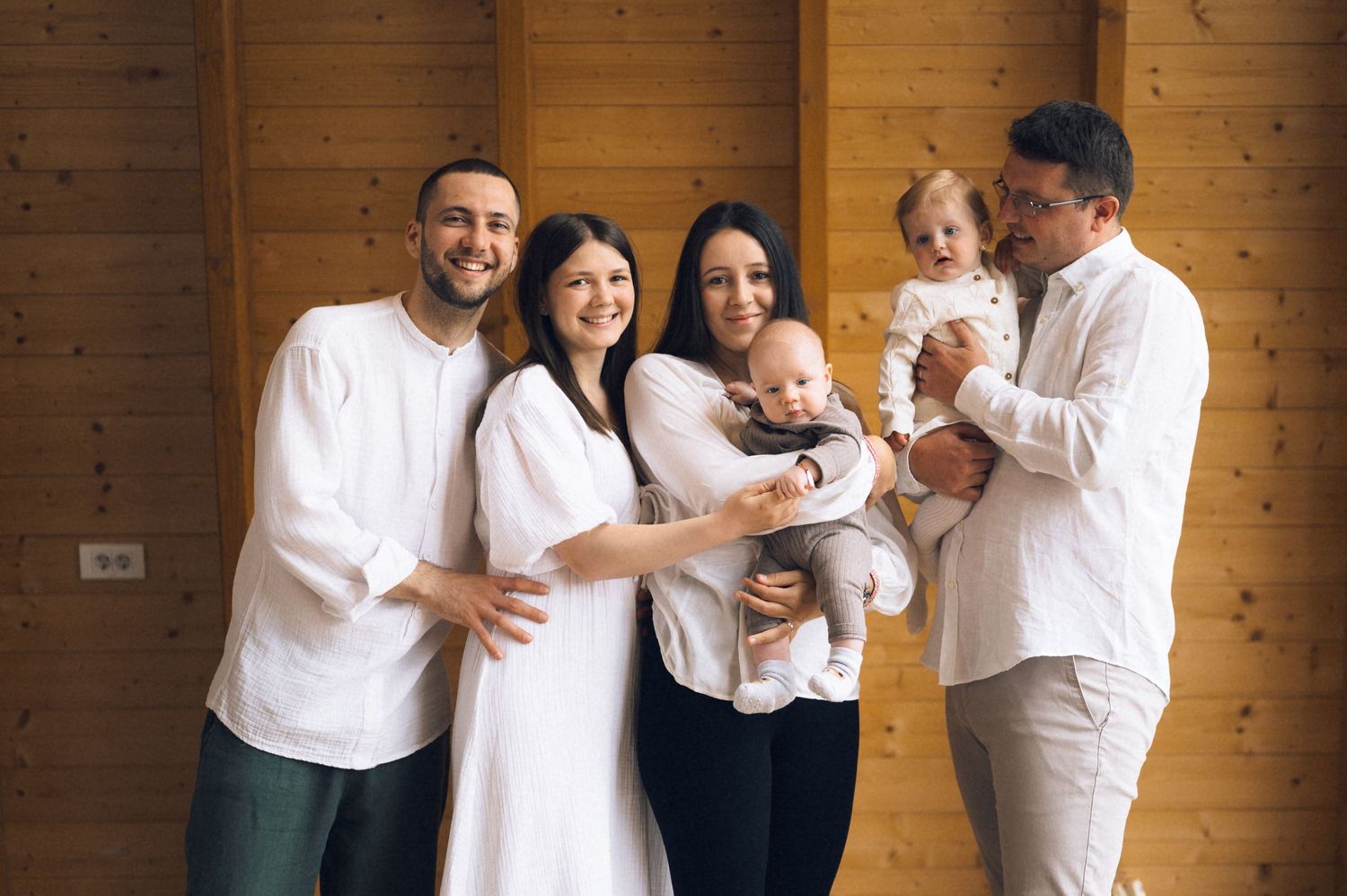 Happy family group of six posing indoors, wearing white outfits, holding two babies, smiling in a wooden room.