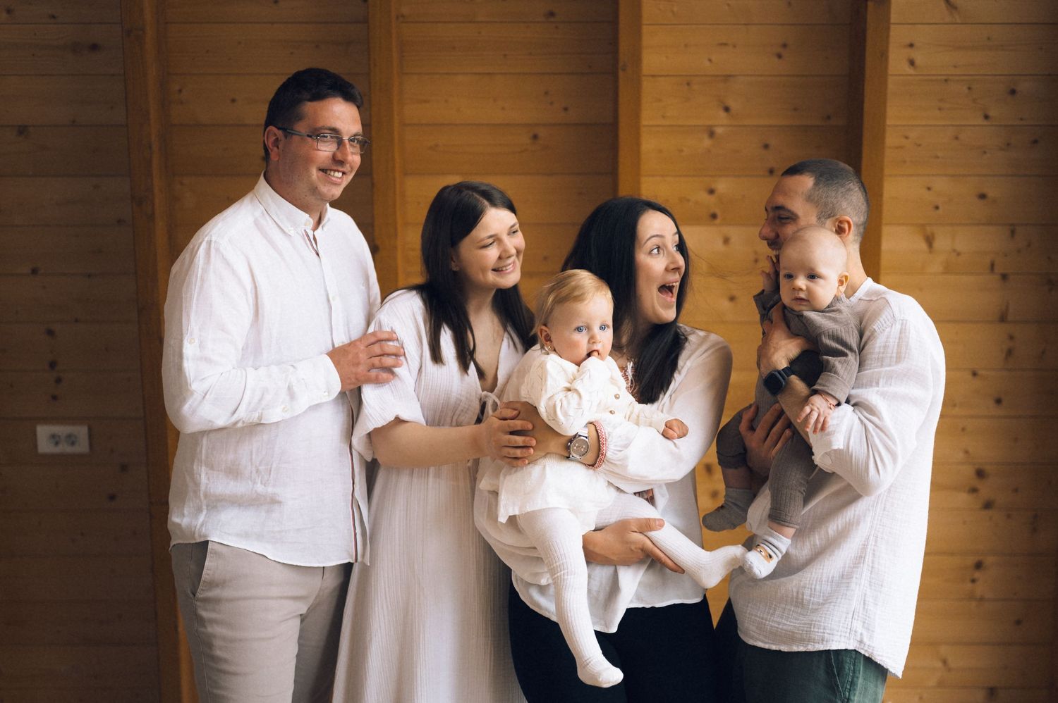 Happy family group portrait with two adults holding babies, smiling indoors against wooden wall background.
