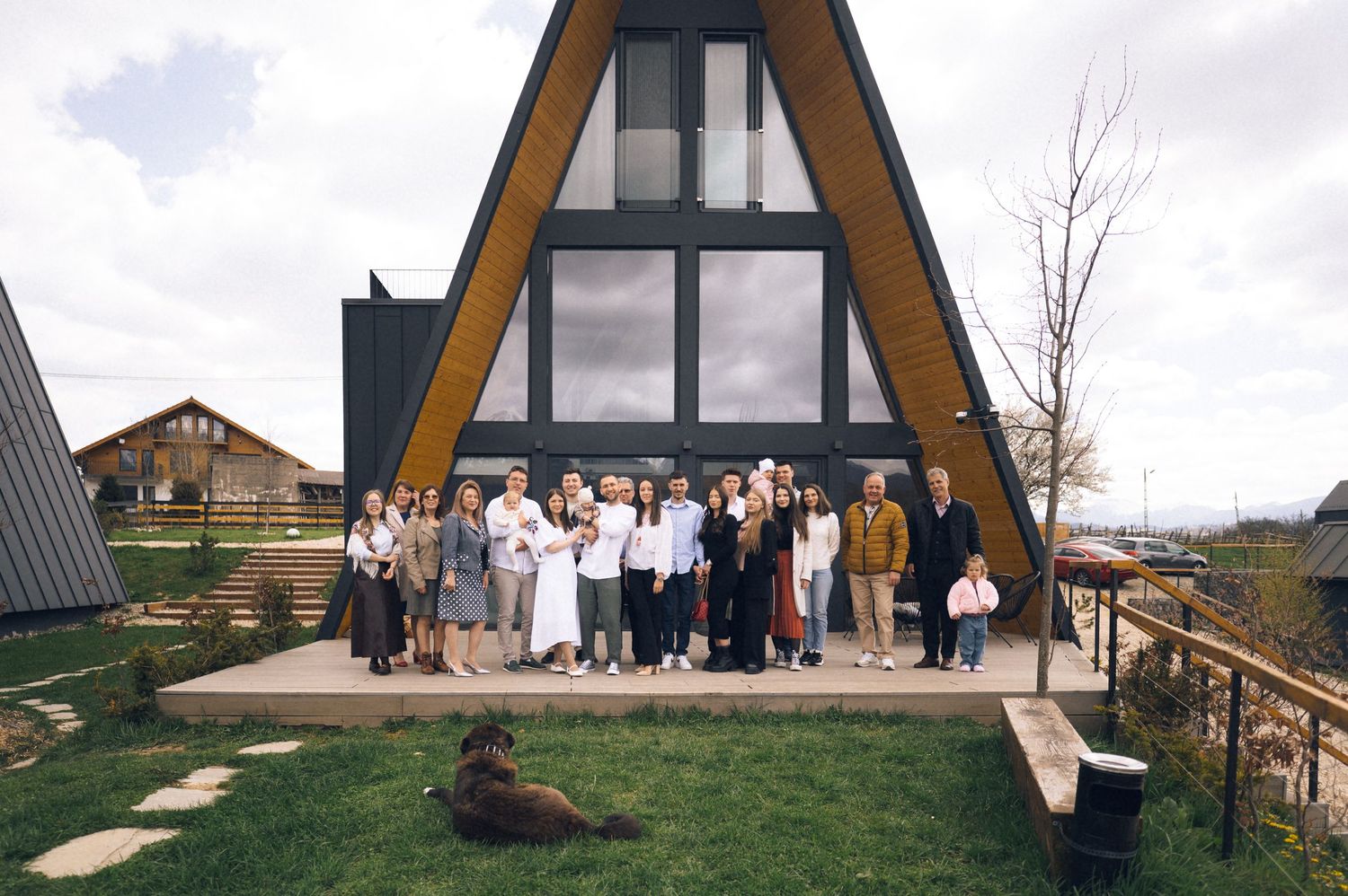 Large group posing in front of modern A-frame cabin with black exterior and yellow accents on green lawn.