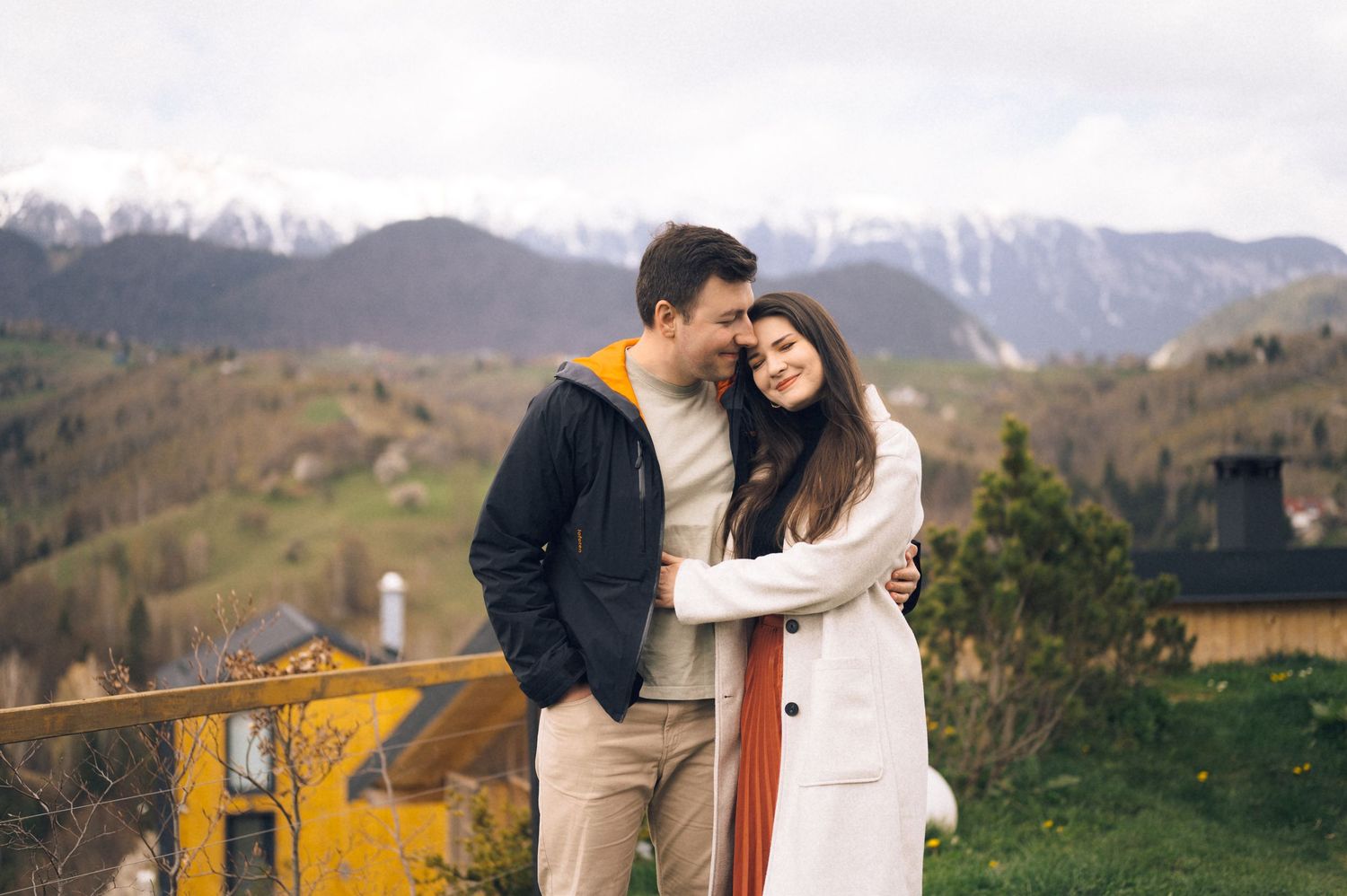 Couple embracing on a balcony with scenic mountain views and green hills in the background.