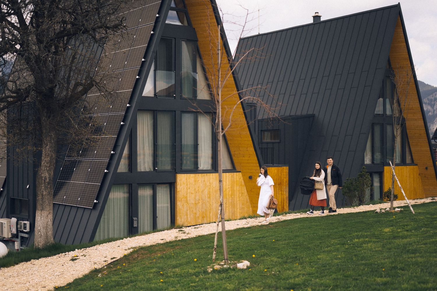 A-frame chalets with black roofs and wooden facades on a green hillside with people walking outside.