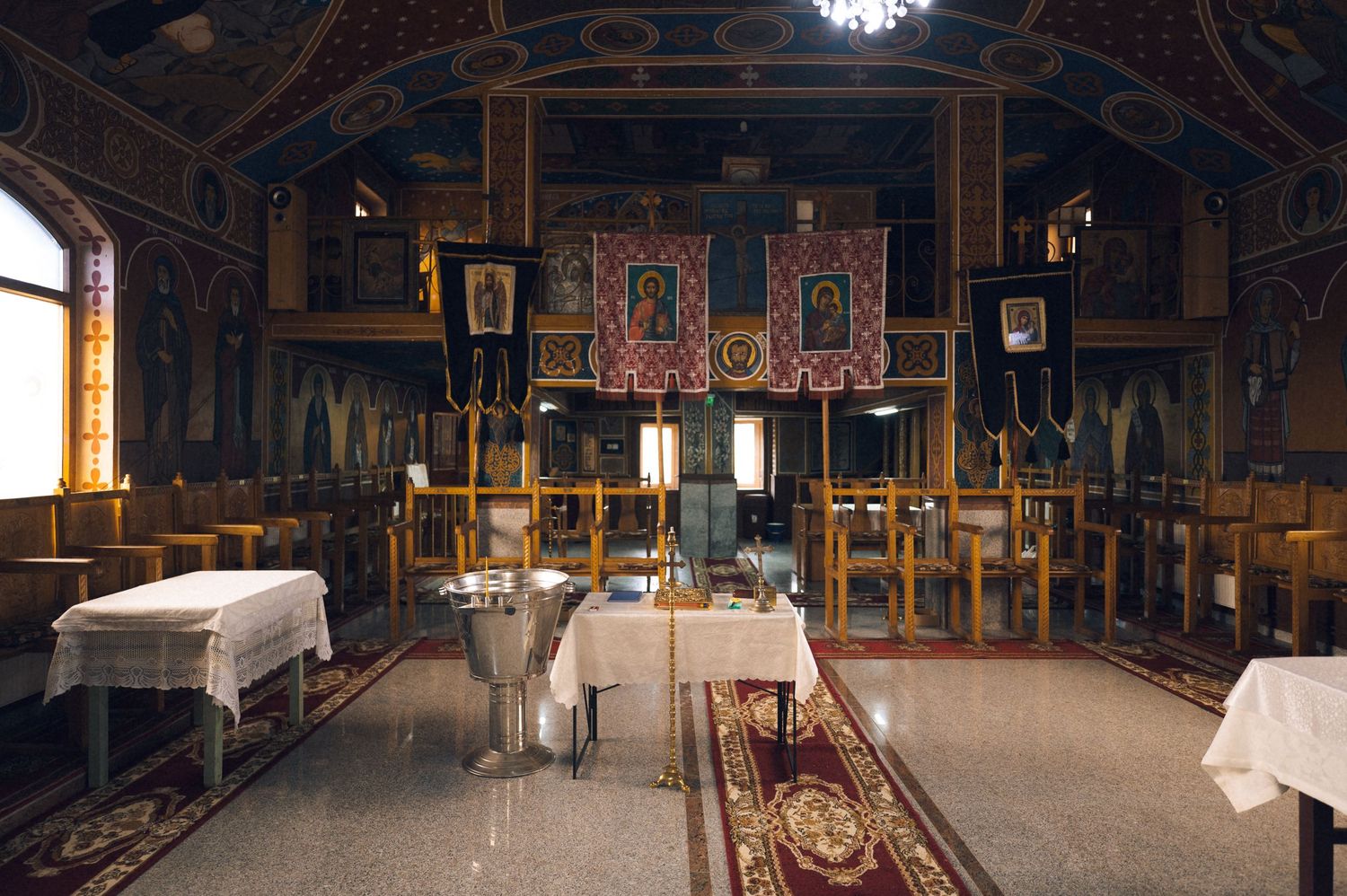 Interior of an ornate Orthodox church with wooden pews, colorful religious icons, and decorative tapestries on the walls.