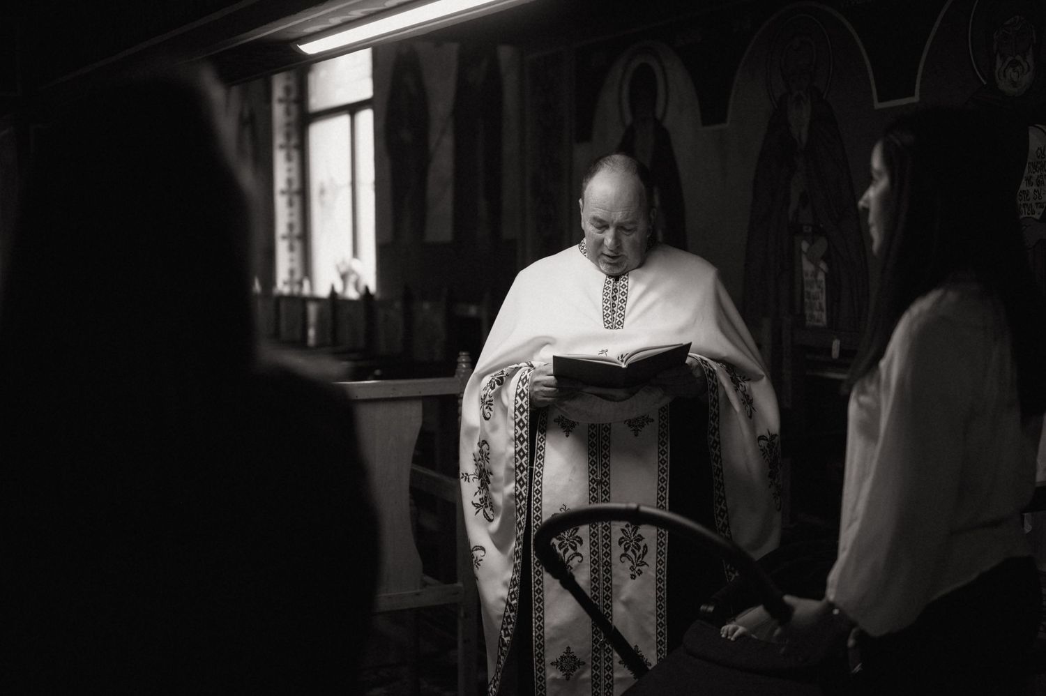 Priest in white vestments reads from a book during a religious ceremony in a dimly lit Orthodox church.
