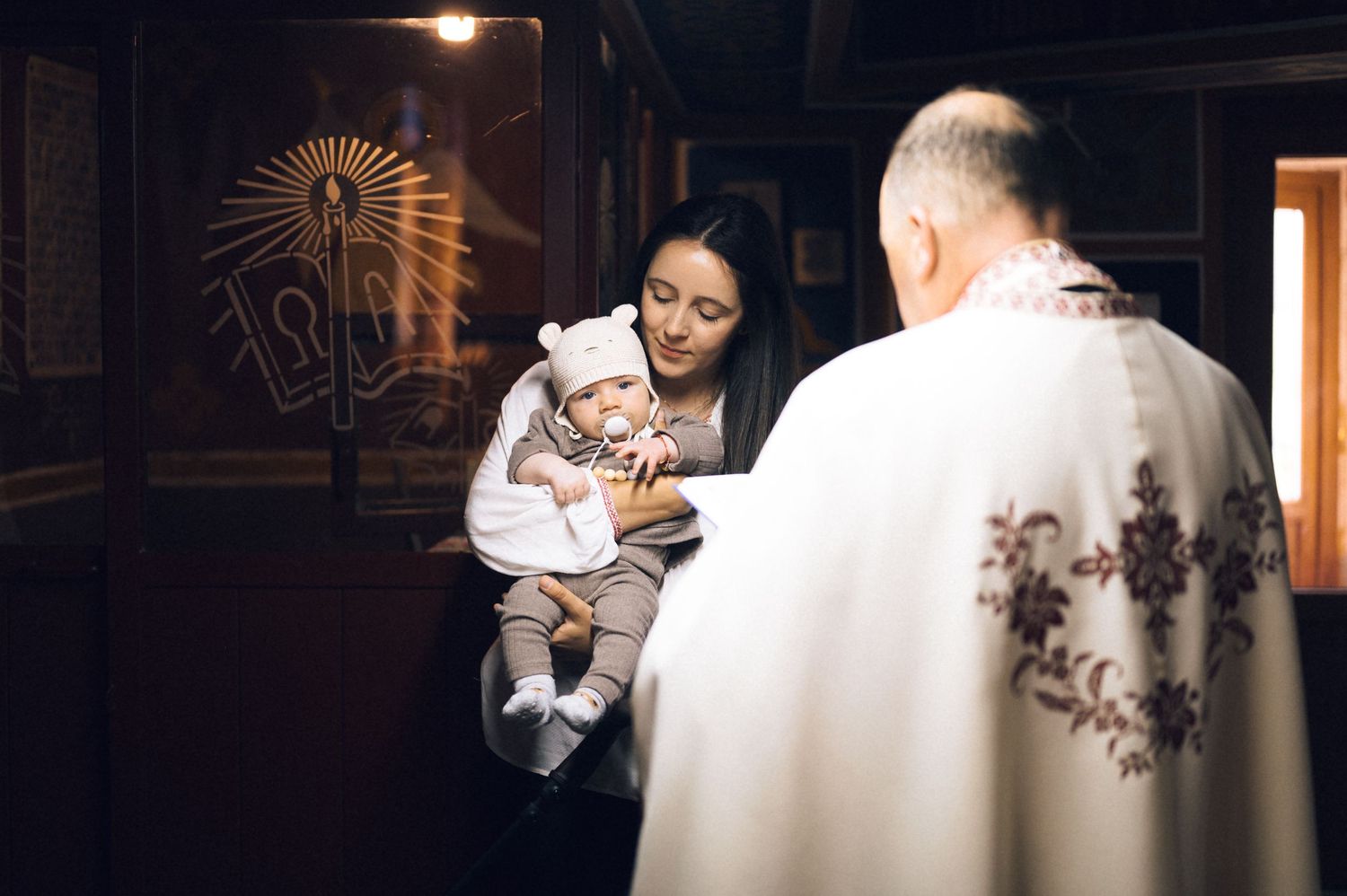 A priest performs a baptism ceremony for a baby in a white bear hat held by its mother in a church.