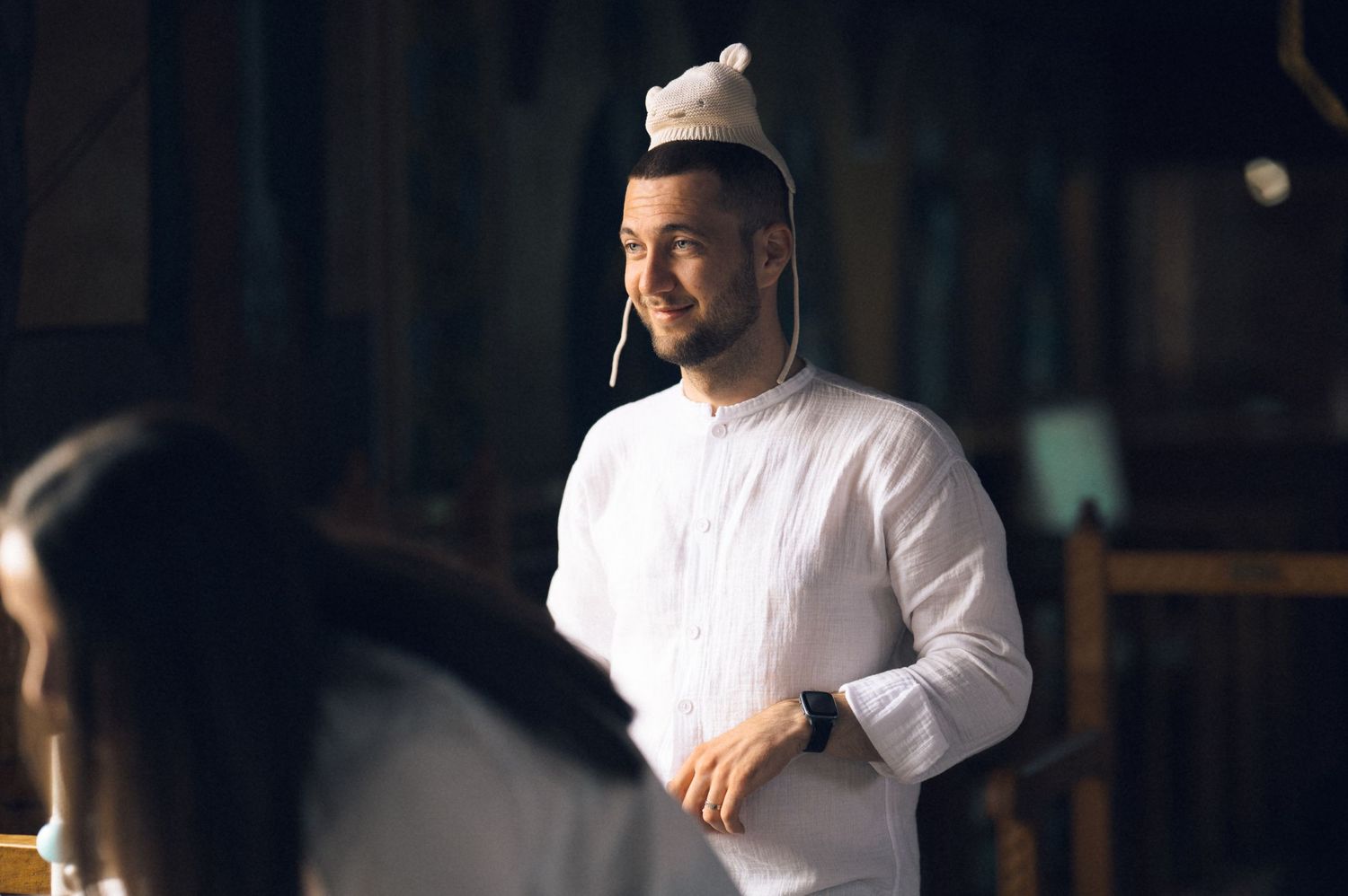 Young man in white traditional attire and kufi cap smiling warmly in a softly lit indoor setting.