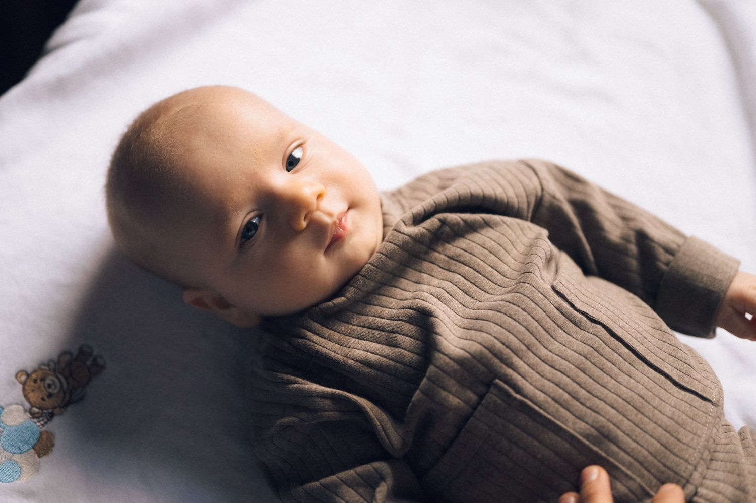 A cute baby in a brown ribbed knit outfit lying on a white blanket, looking up with bright curious eyes.