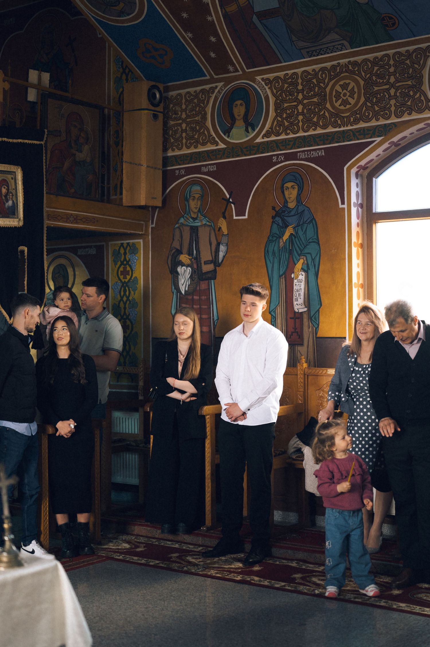 Family gathering inside an Orthodox church with colorful Byzantine icons and murals on the walls during a religious ceremony.