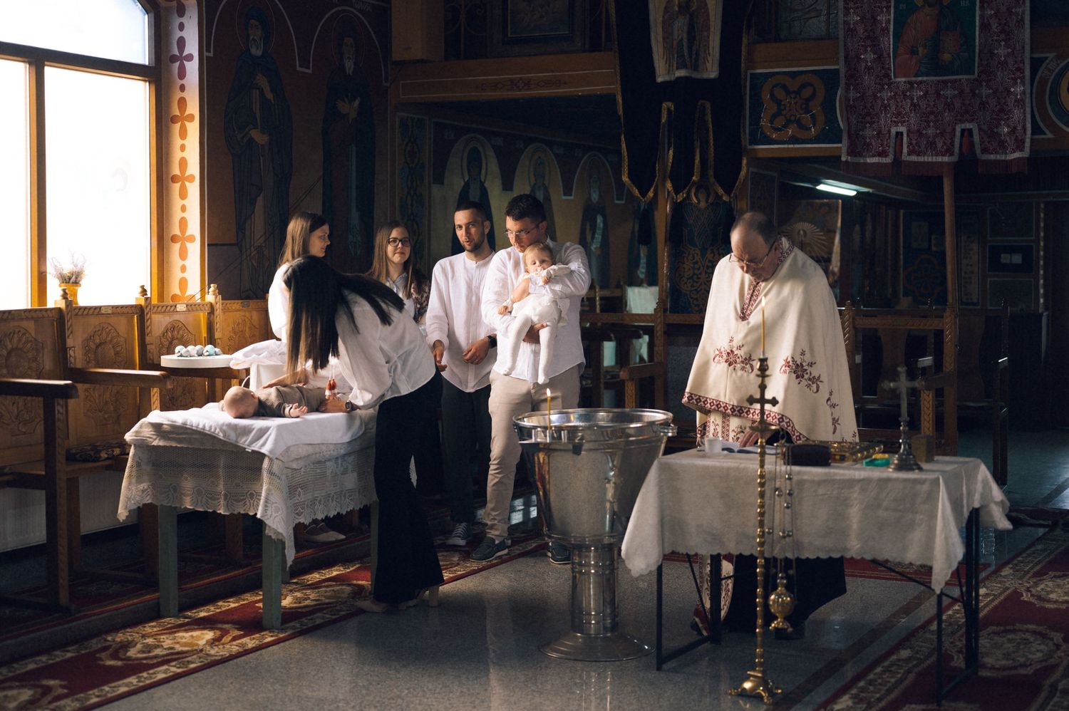 Orthodox Christian baptism ceremony with priest and family gathered around a baptismal font in a church.