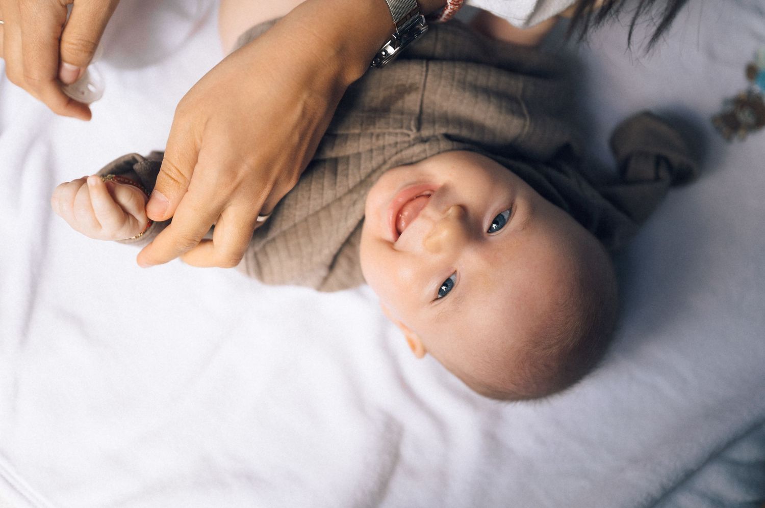 Smiling baby in brown outfit lying on white blanket while adult hands gently hold them during diaper change.
