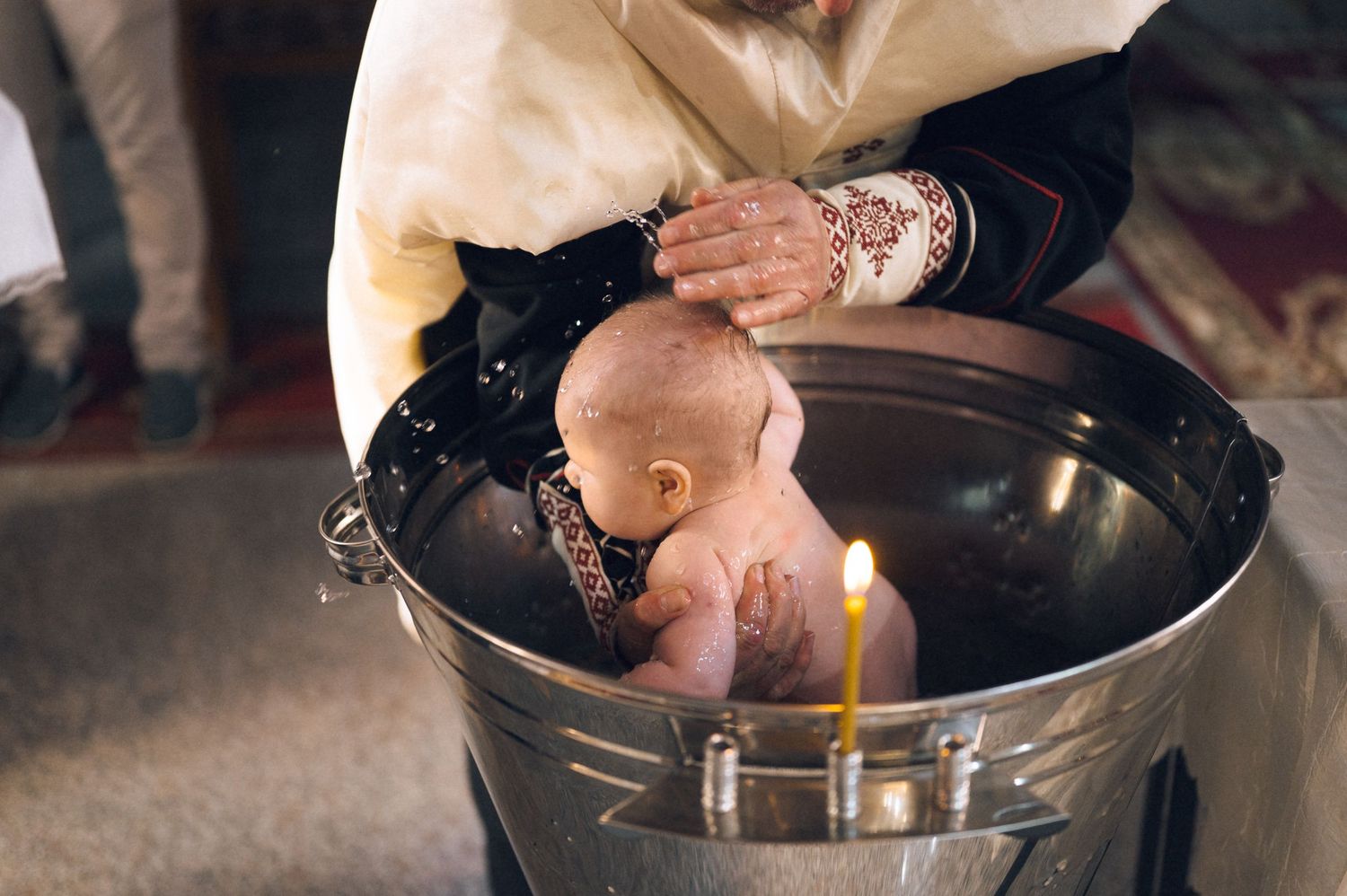 Baby being baptized in a metal bucket with a lit candle during a traditional Orthodox Christian baptism ceremony.