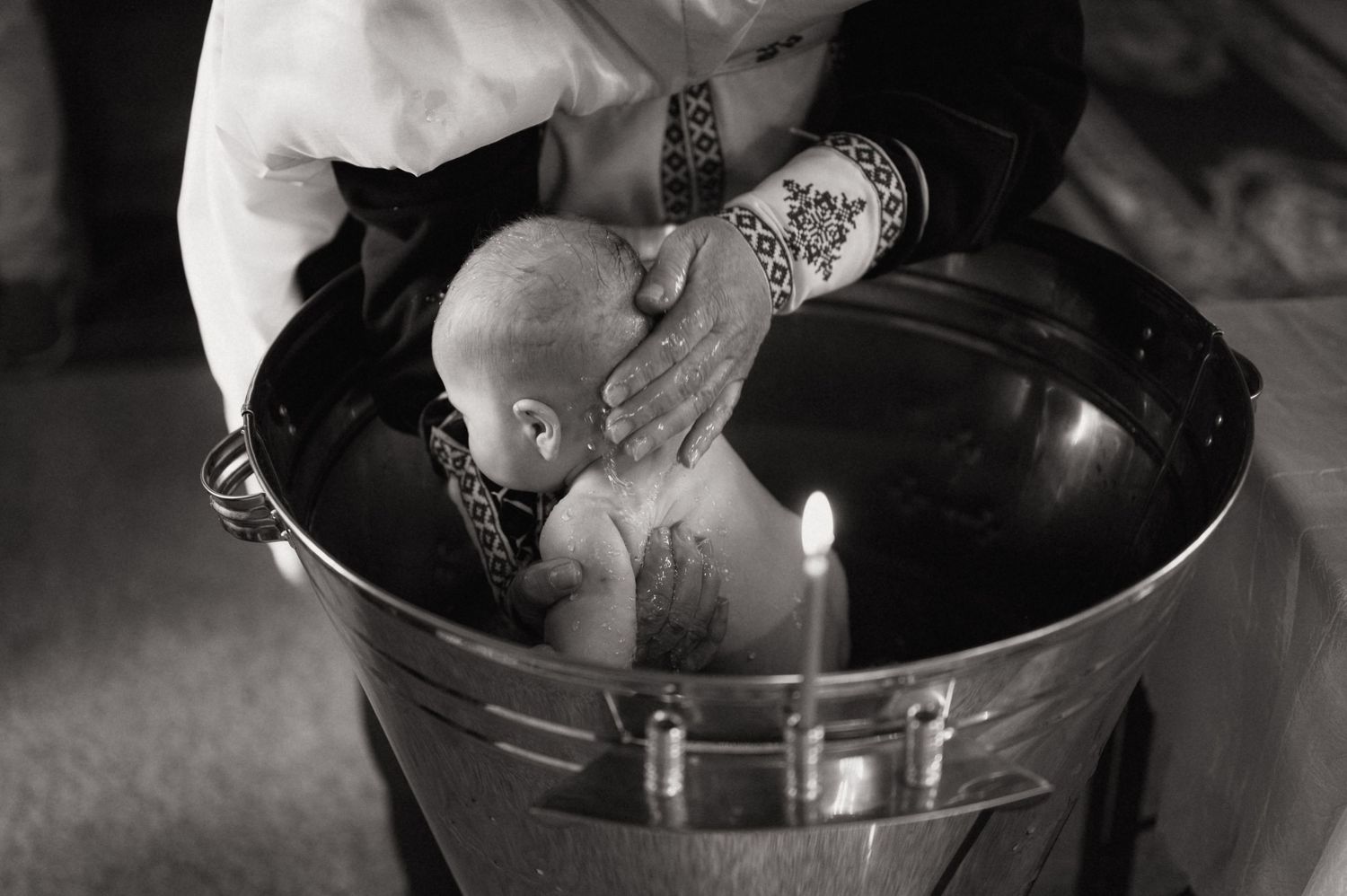 Black and white photo of a baby being baptized in a metal font with a lit candle nearby.