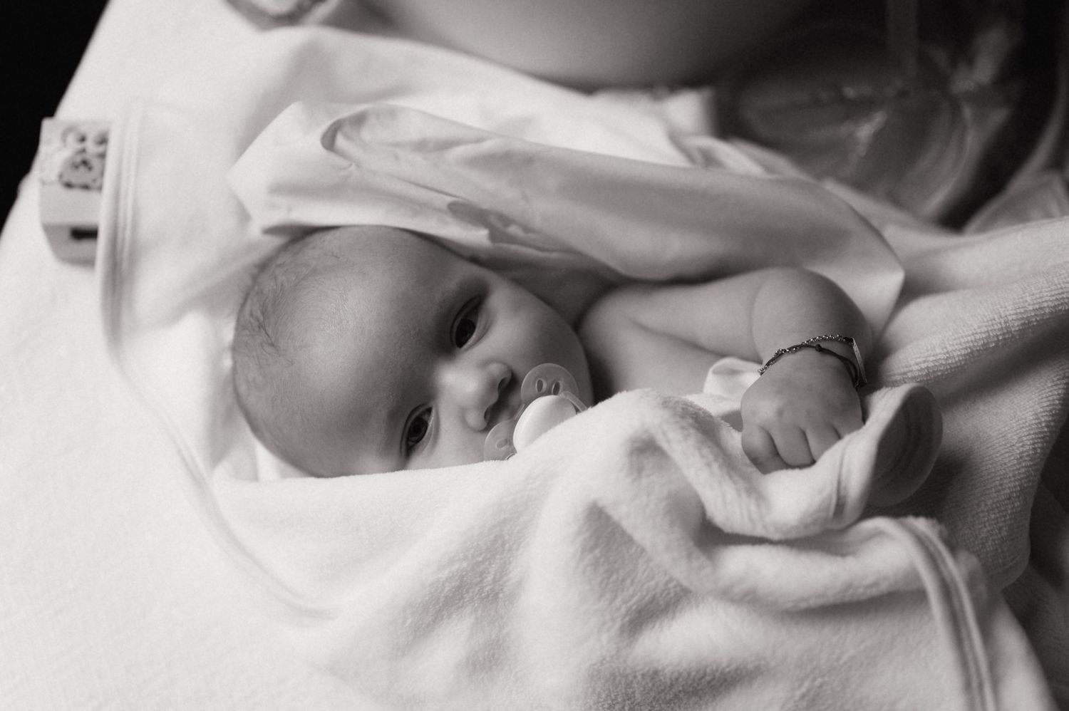 Black and white photo of a peaceful newborn baby wrapped in a soft white blanket with a pacifier.