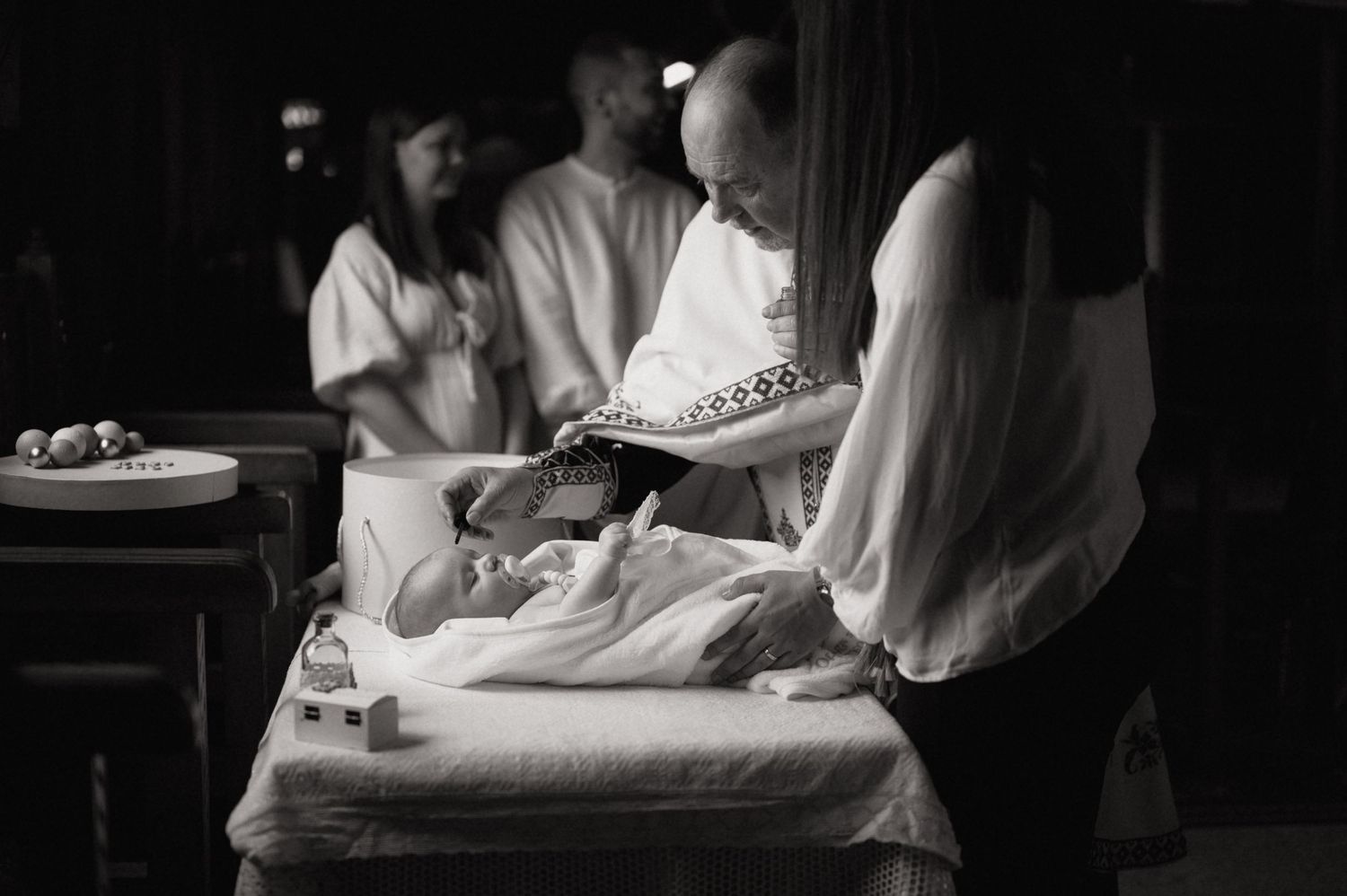 Black and white photo of a priest baptizing a newborn baby with family members gathered around.