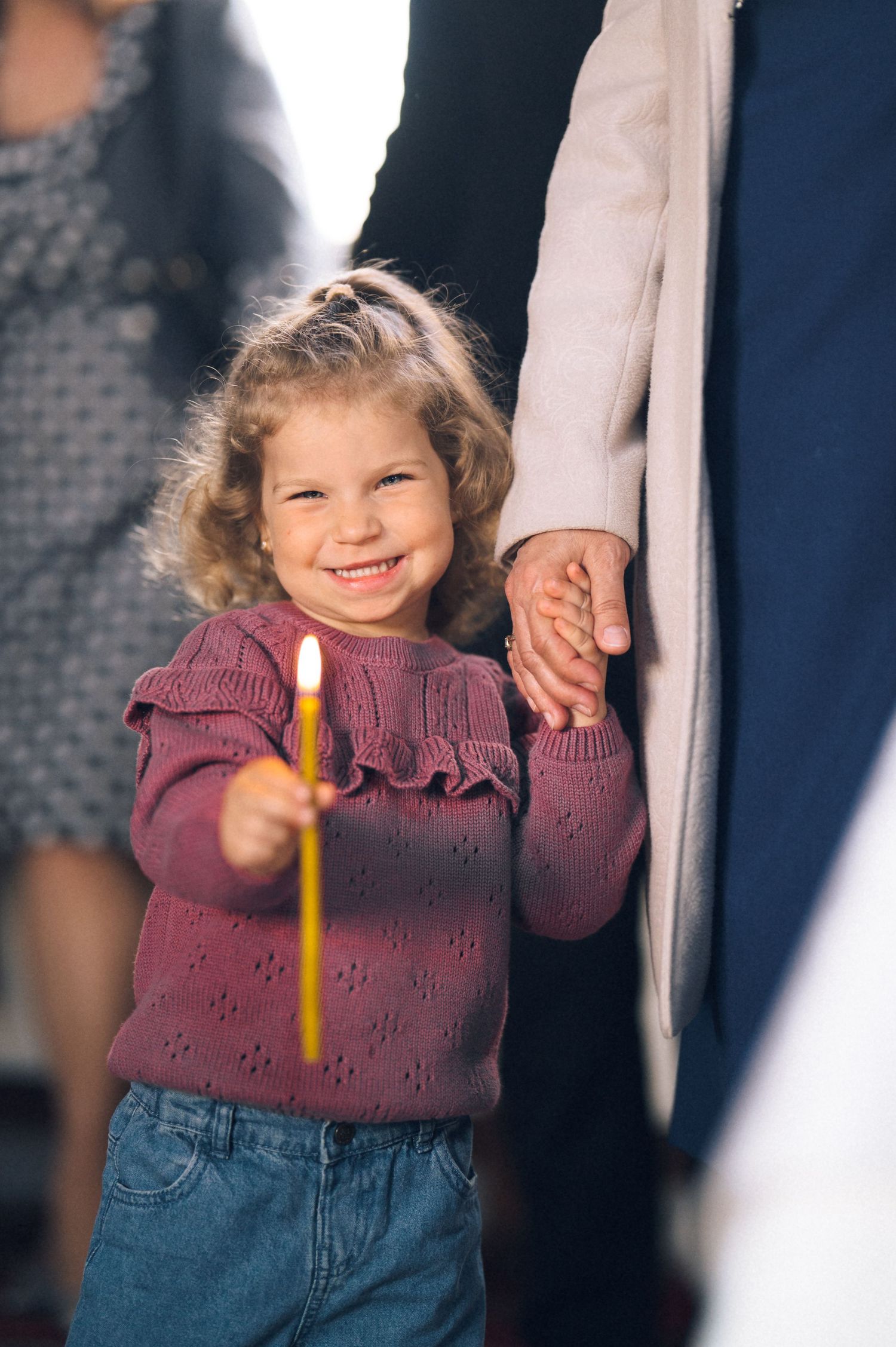 Smiling little girl in purple sweater holding a candle and an adult's hand outdoors.