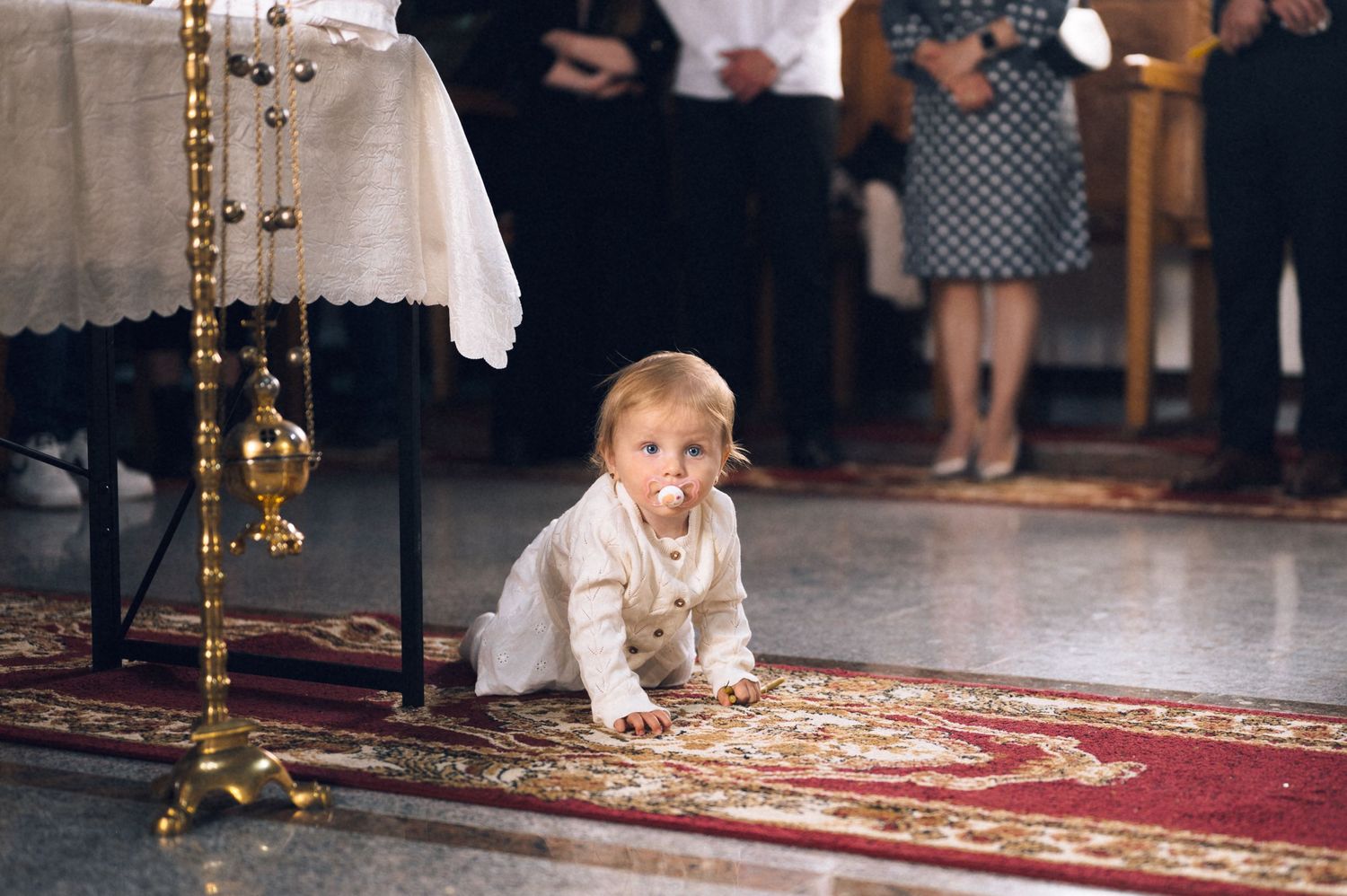 Baby in white outfit crawling on ornate red carpet inside a church during a baptism ceremony.