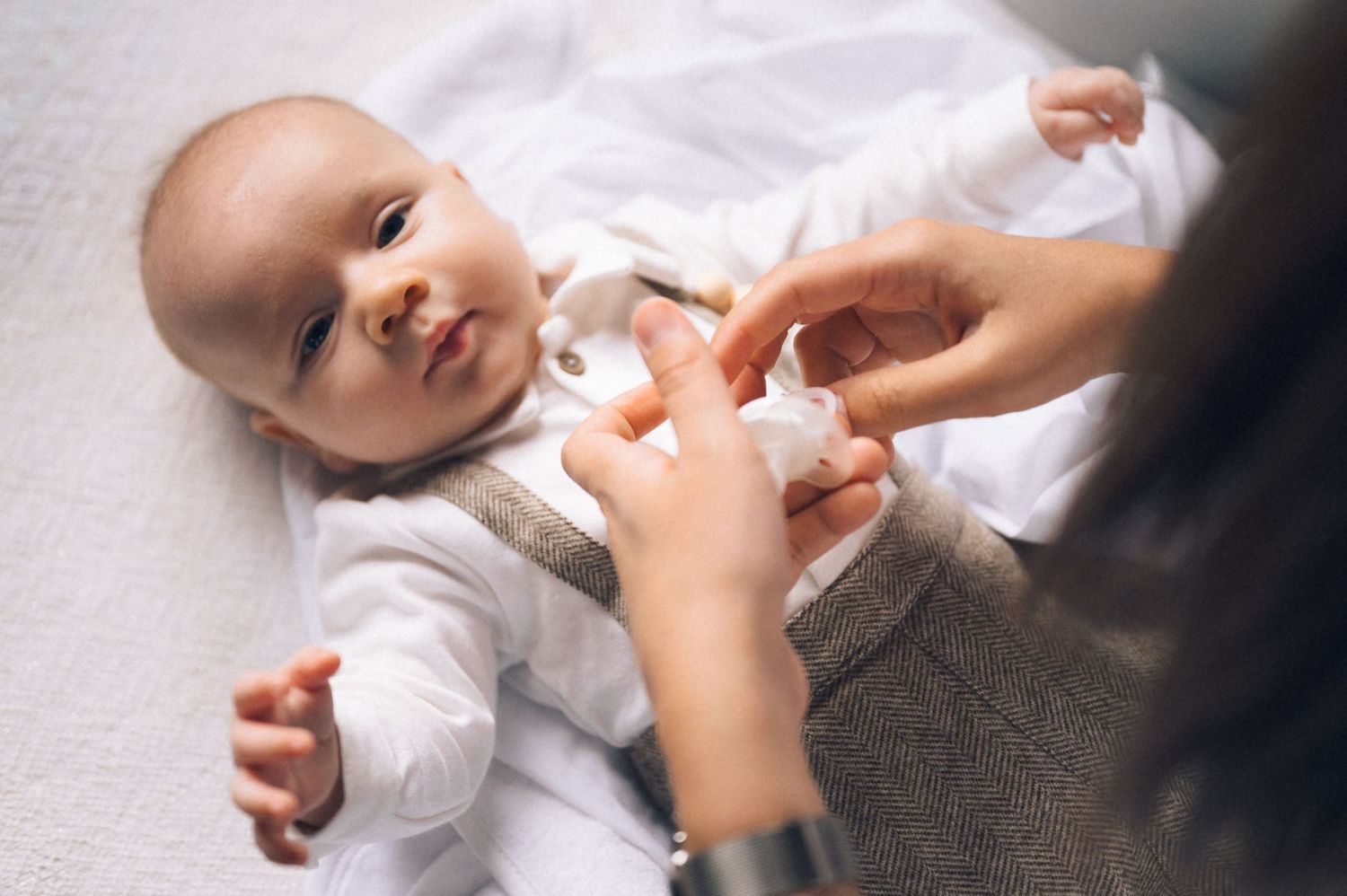 Mother gently cleaning her smiling baby lying on a white surface, wearing a white onesie.