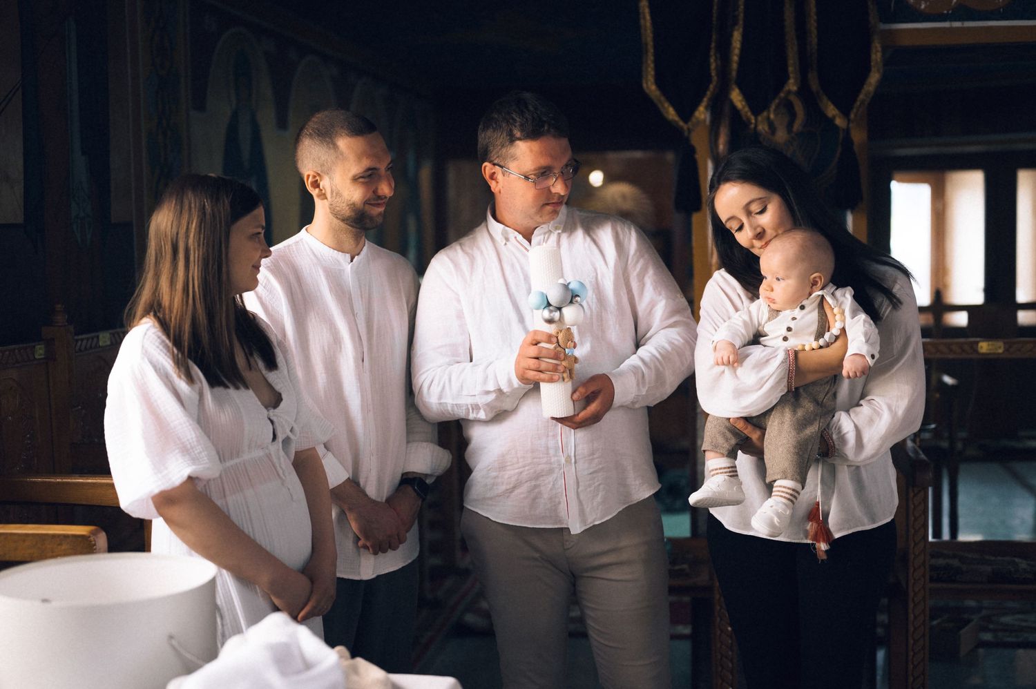 Family gathered in a church for a baptism ceremony, dressed in white, with parents holding a newborn baby.