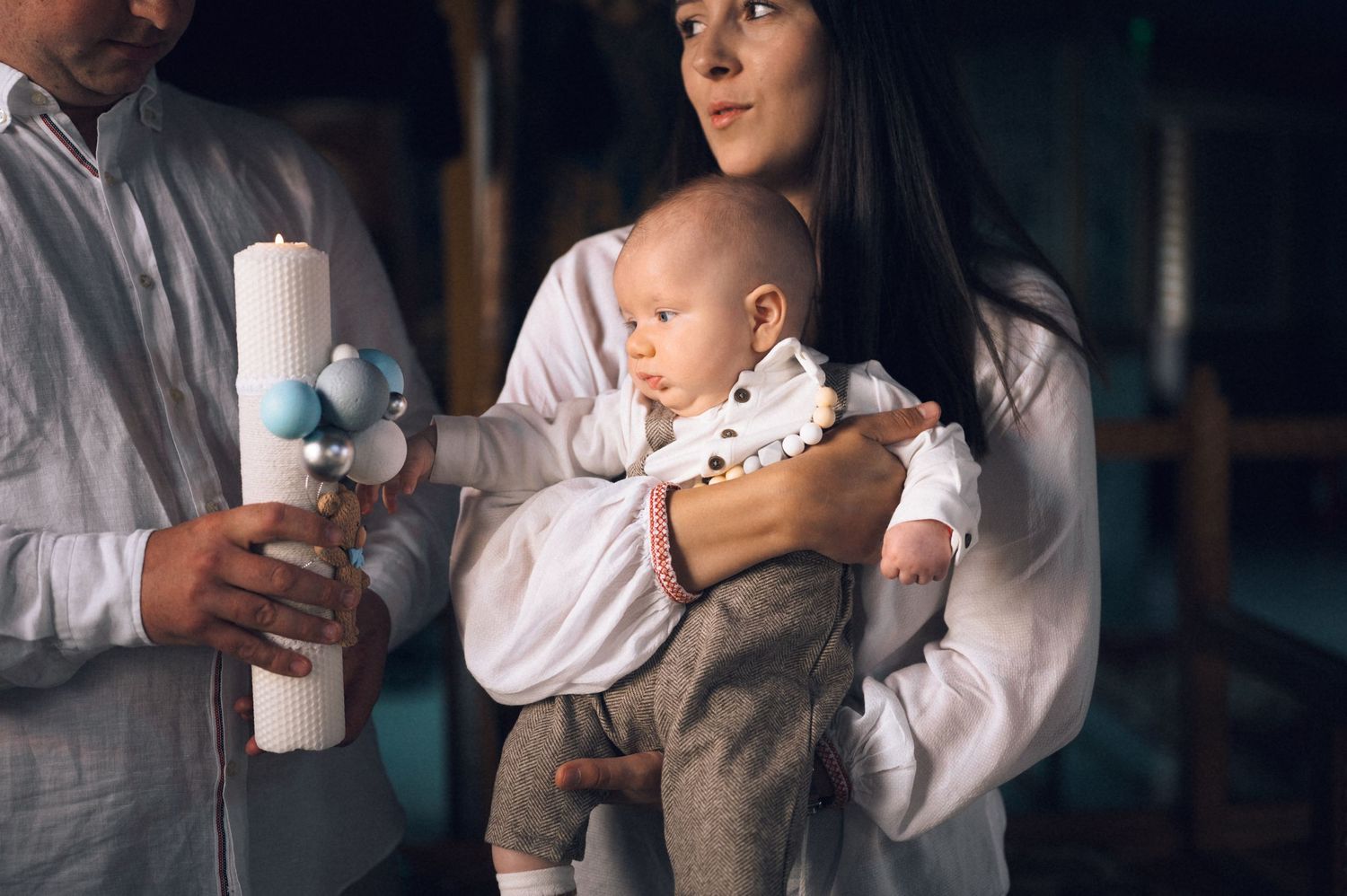 A woman in white holds a baby at a christening ceremony while a man holds a decorative baptismal candle.