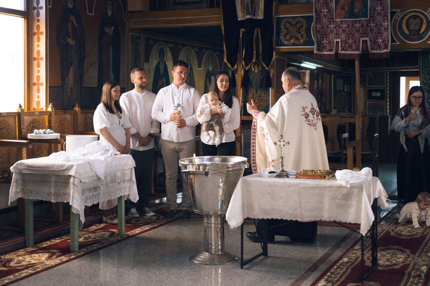 Orthodox Christian baptism ceremony with priest and godparents gathered around a baptismal font inside a wooden church.