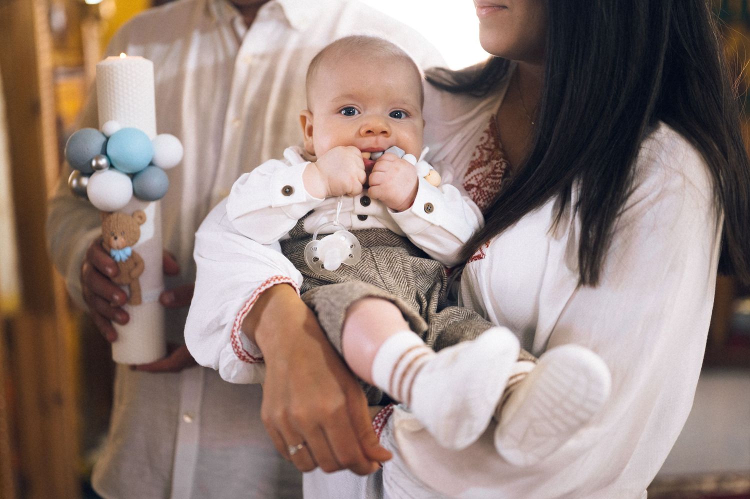 Parents holding a smiling baby in white outfit at a christening ceremony with a decorative baptismal candle.