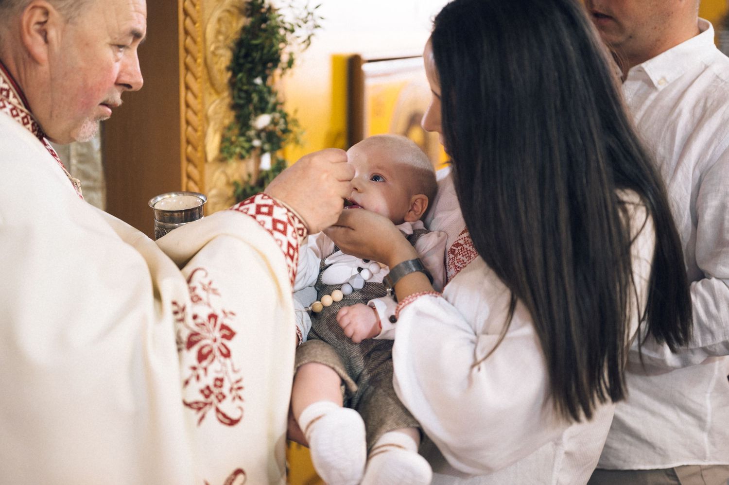 Priest performing Catholic baptism ceremony on infant held by mother in church.