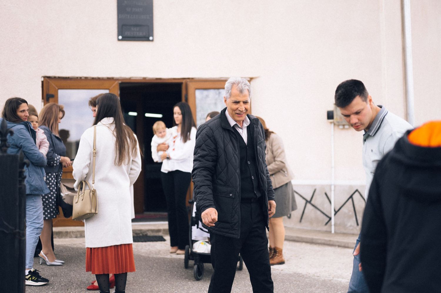 Group of people gathered outside a white building, man in black jacket walking in foreground.