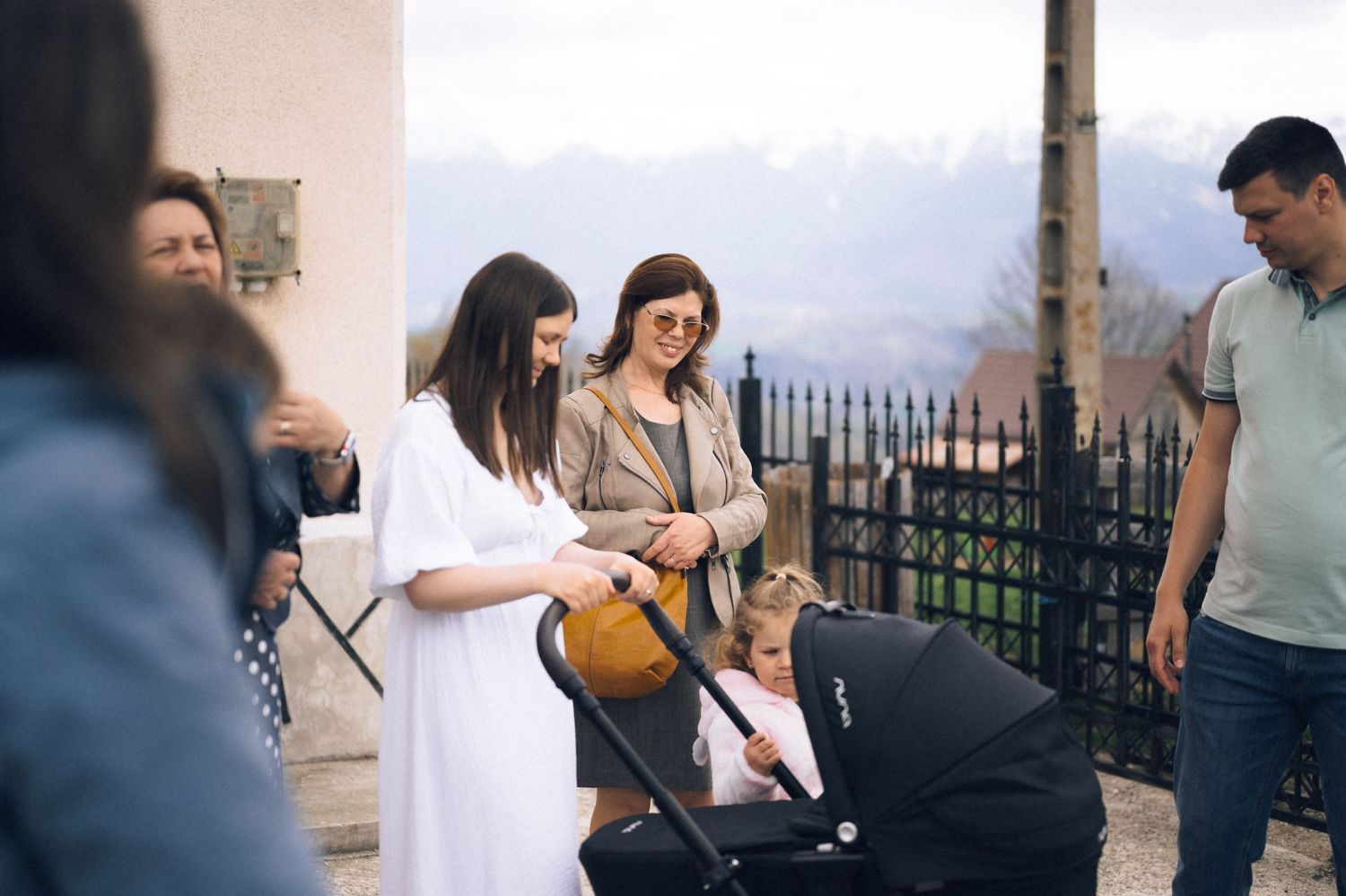 Two women with a baby stroller chat outdoors with mountain views, joined by a man in a casual setting.