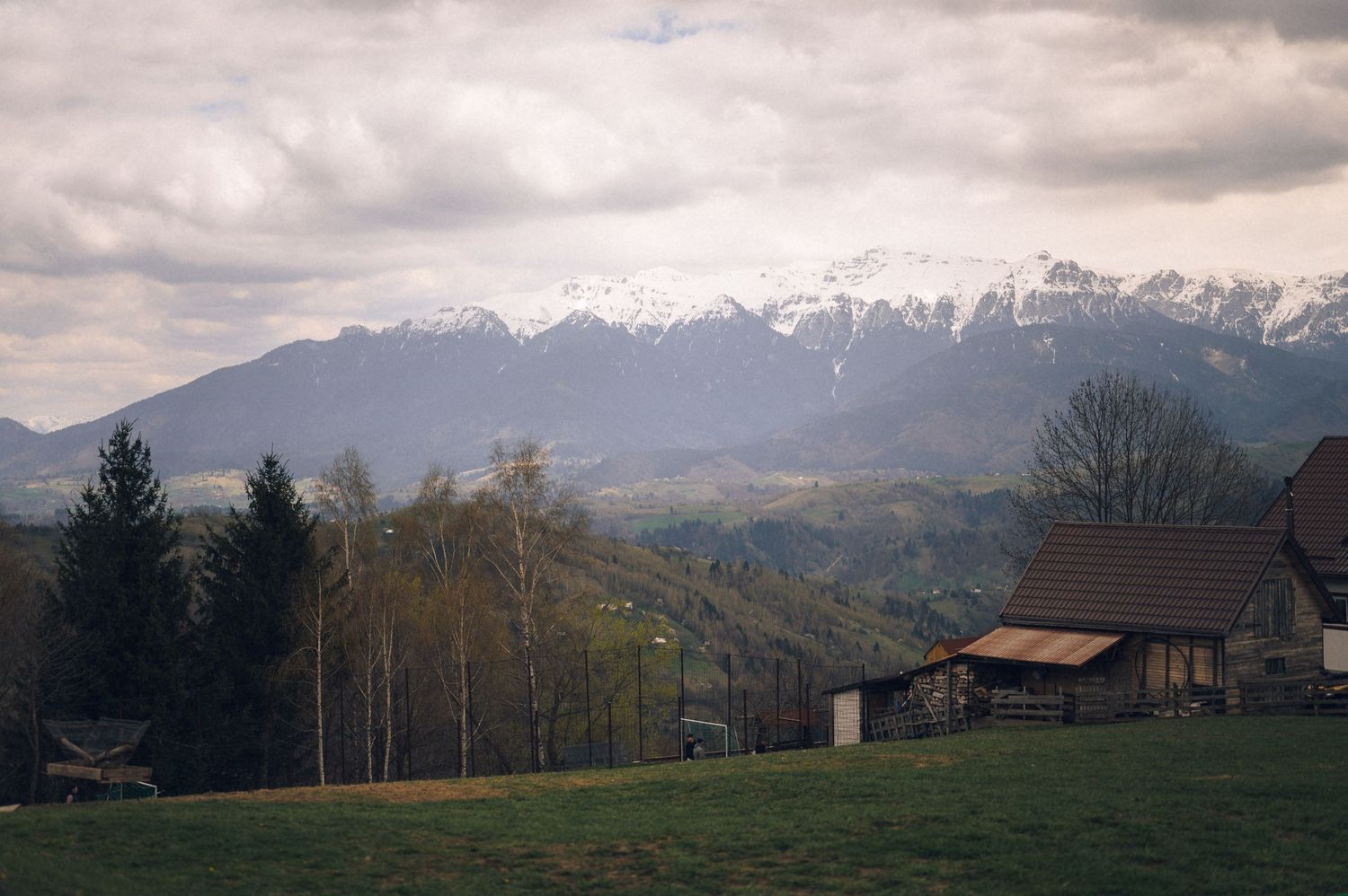 Snow-capped mountain range viewed from a green hillside with a rustic wooden house and trees under cloudy skies.