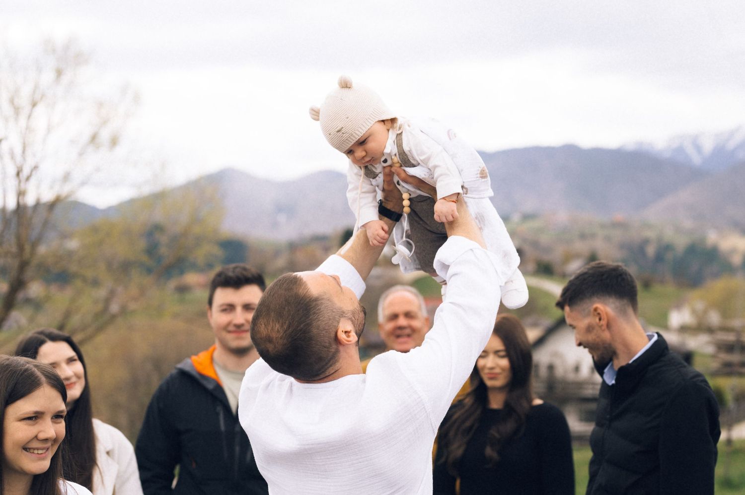 Man lifting a baby in the air outdoors, surrounded by smiling family members with mountains in the background.