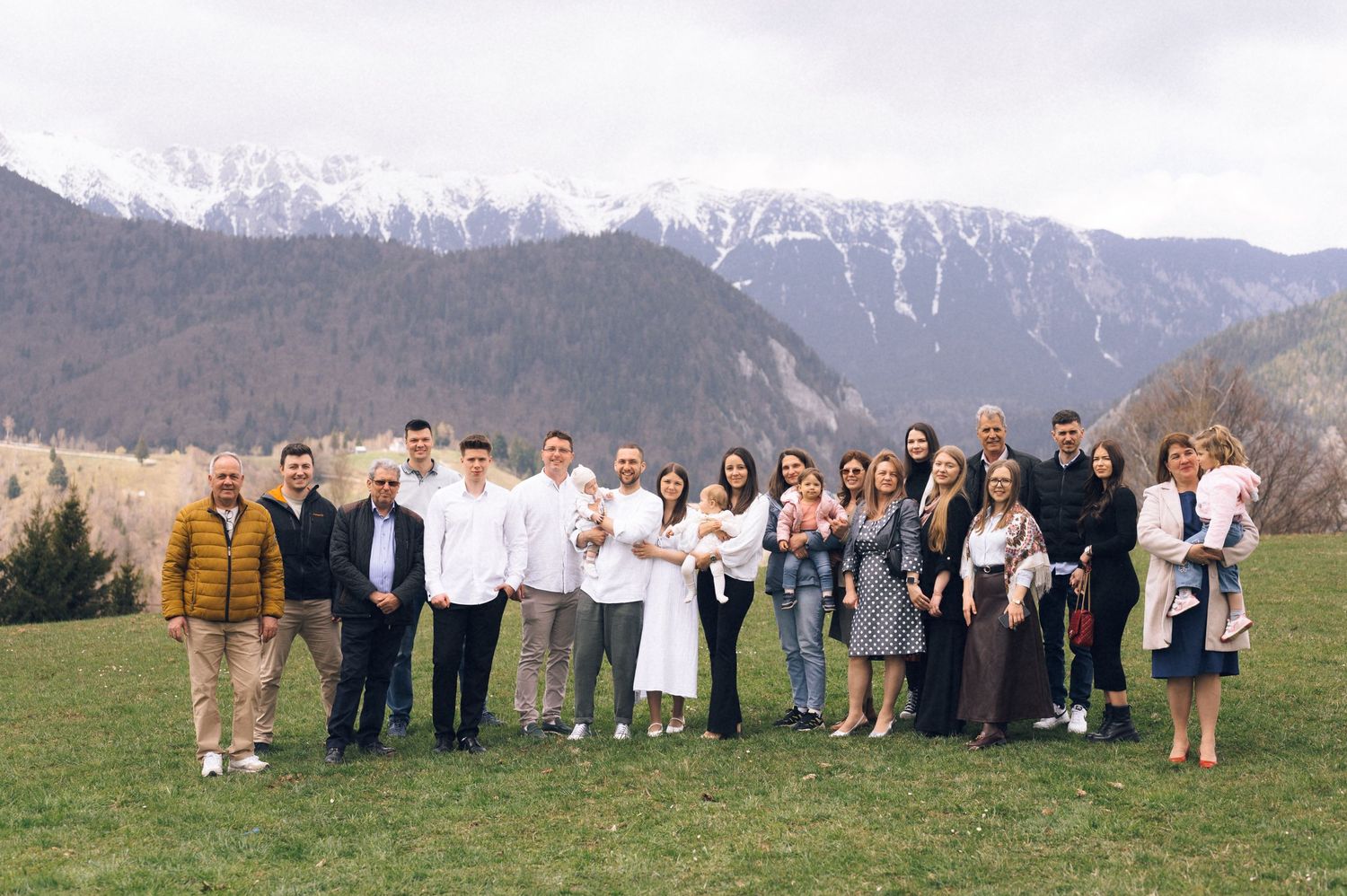 Large family group photo outdoors with snow-capped mountains and green meadow backdrop.