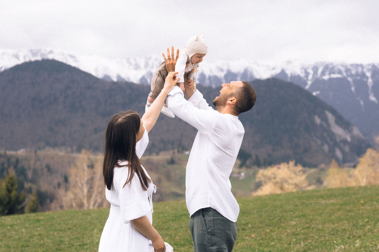 Happy family with baby enjoying mountain meadow, snow-capped peaks in background, parents lifting infant joyfully.