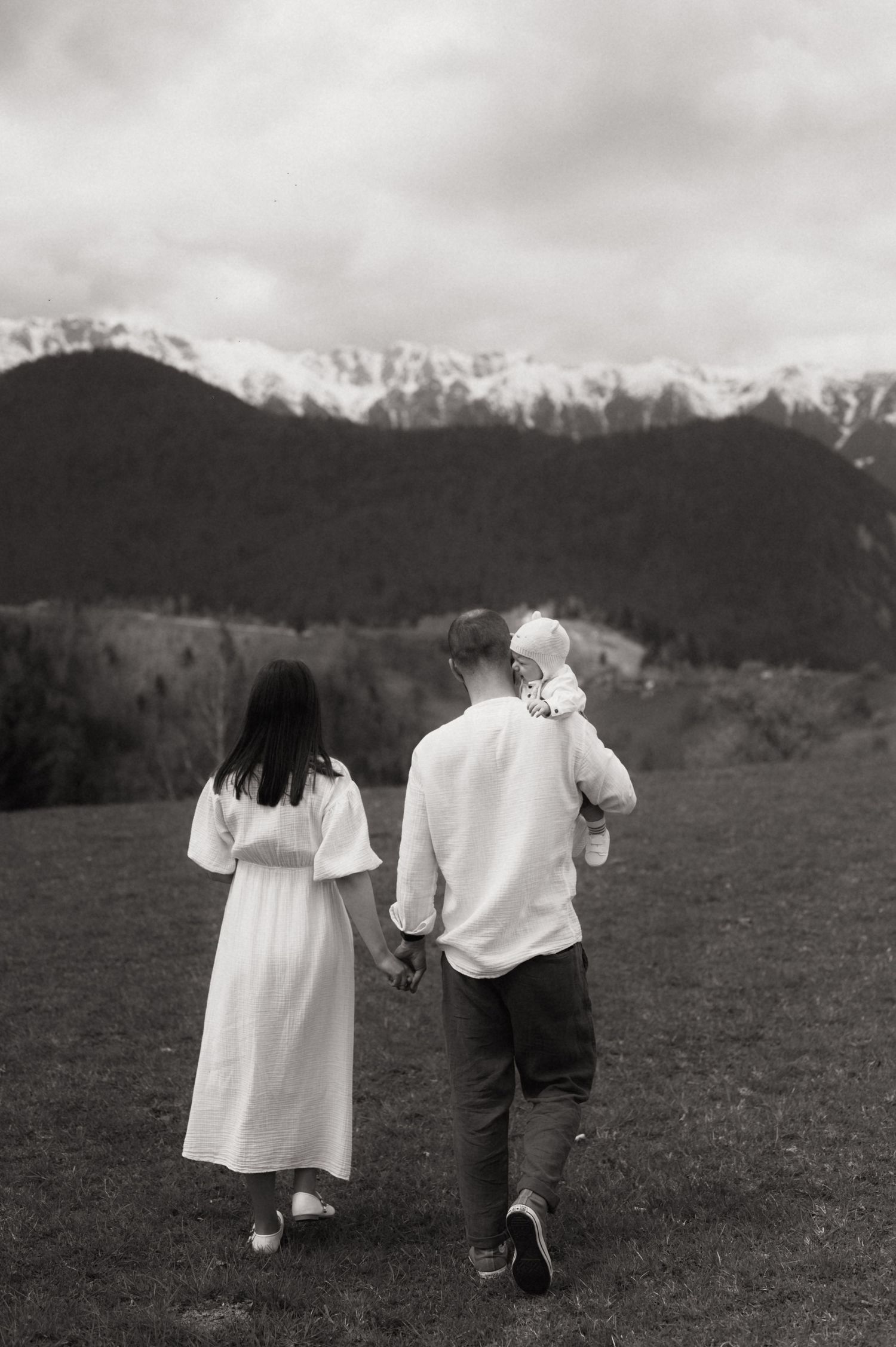 Black and white photo of a family walking in a mountain meadow, parents holding hands with father carrying a baby.