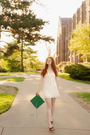 Young redhead woman in white dress holding green graduation cap on a sunny college campus pathway.