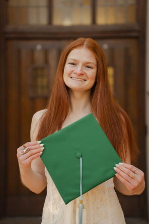 Smiling redhead woman holding a green graduation cap in front of wooden doors.